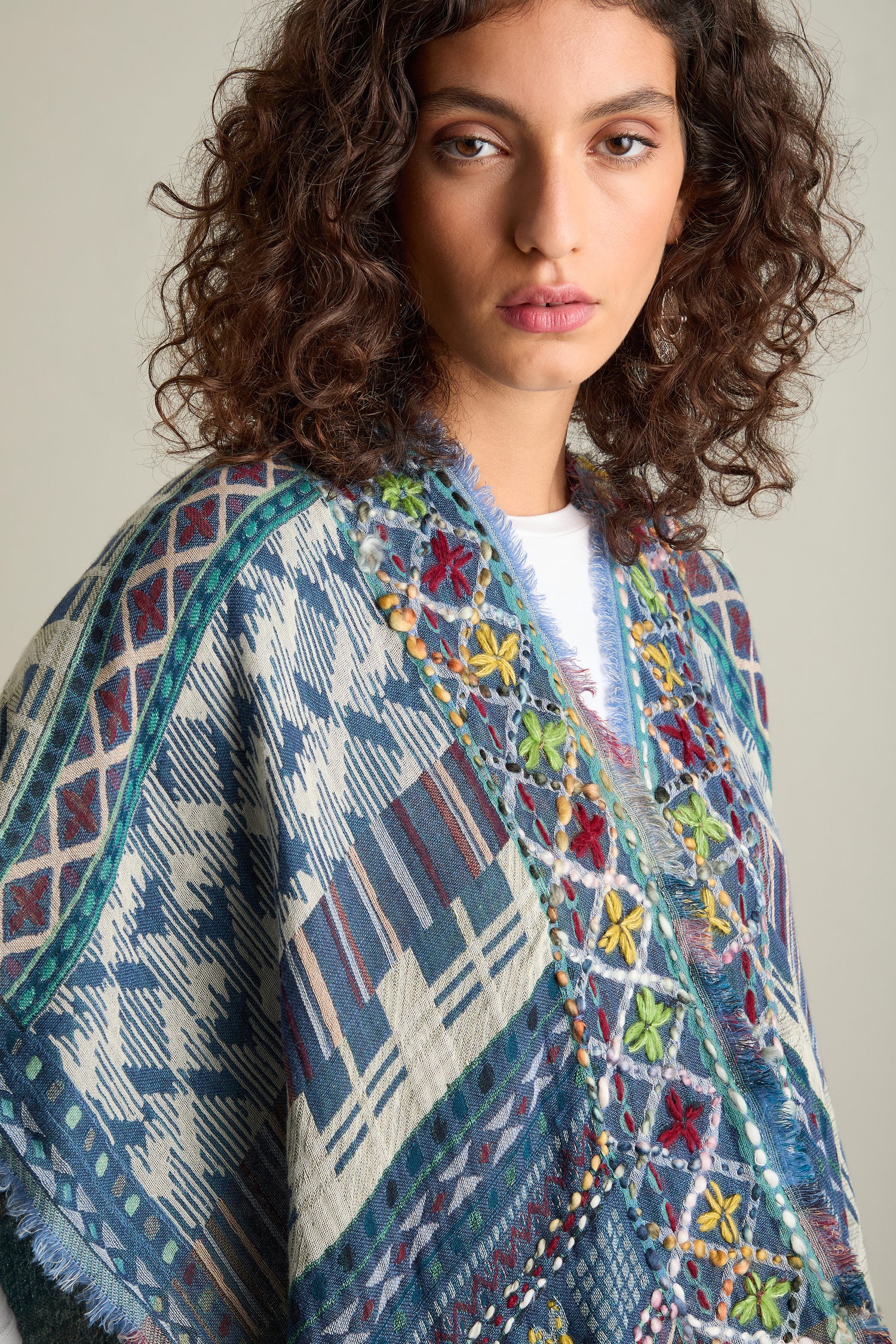 A person with curly hair wears the Embroidered Cotton Shawl in blue and white, adorned with colorful flowers, making it a luxurious accessory.