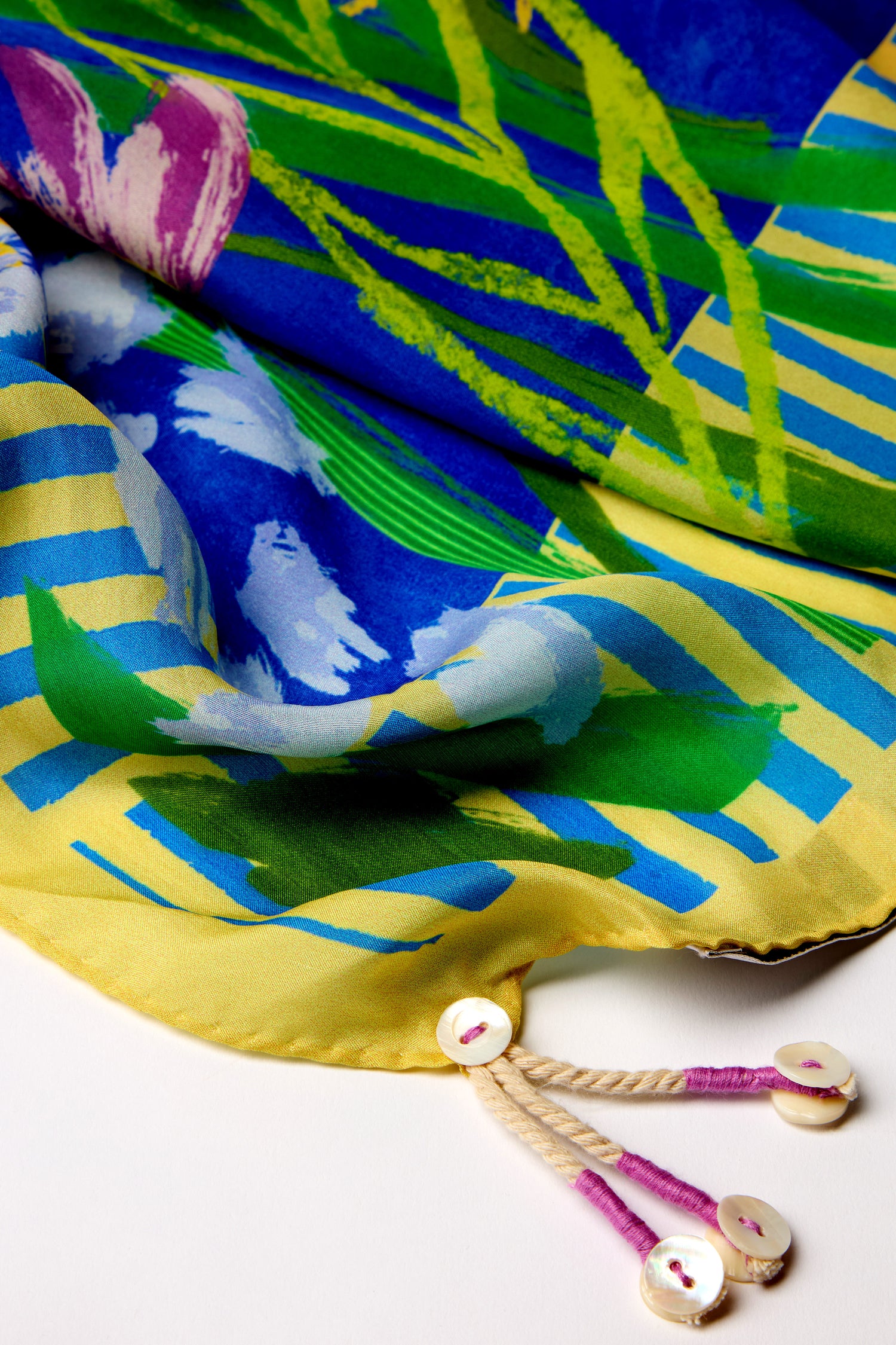 The Garden Paradise Silk Square Scarf showcases a vibrant tropical design with garden-inspired prints in green, blue, and yellow. It features two white buttons attached to beige cords on the edge for added flair.