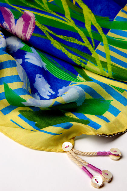 The Garden Paradise Silk Square Scarf showcases a vibrant tropical design with garden-inspired prints in green, blue, and yellow. It features two white buttons attached to beige cords on the edge for added flair.