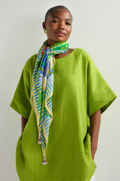 Against a plain light background, someone wearing a green tunic showcases the Garden Paradise Silk Square Scarf adorned with garden-inspired prints, adding vibrant accessories that enhance their ensemble.