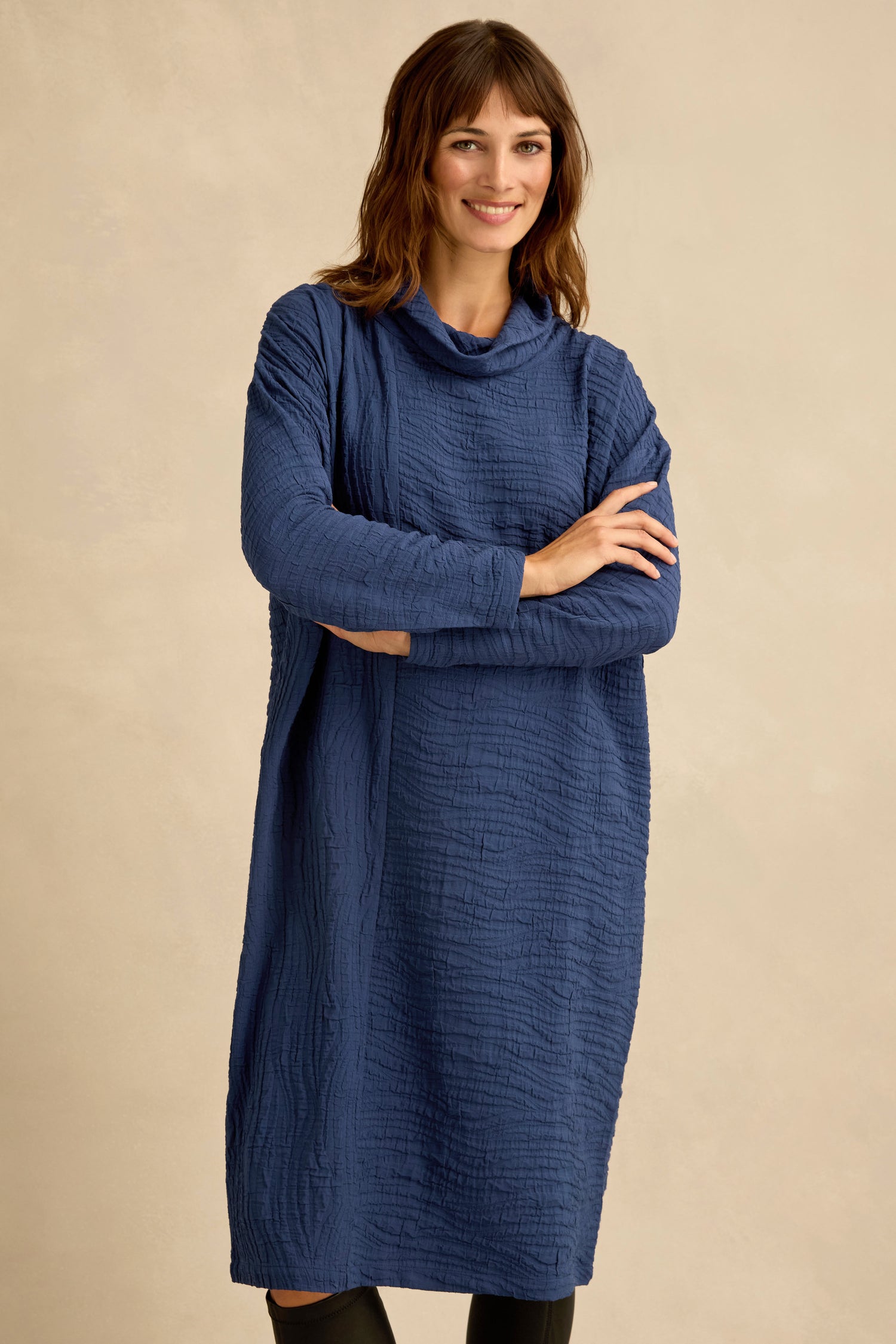 A woman stands with arms crossed, wearing the Wave Textured Crinkle Dress—a loose-fitting, long-sleeved dress in textured navy blue with a subtle bubble silhouette—set against a neutral background.
