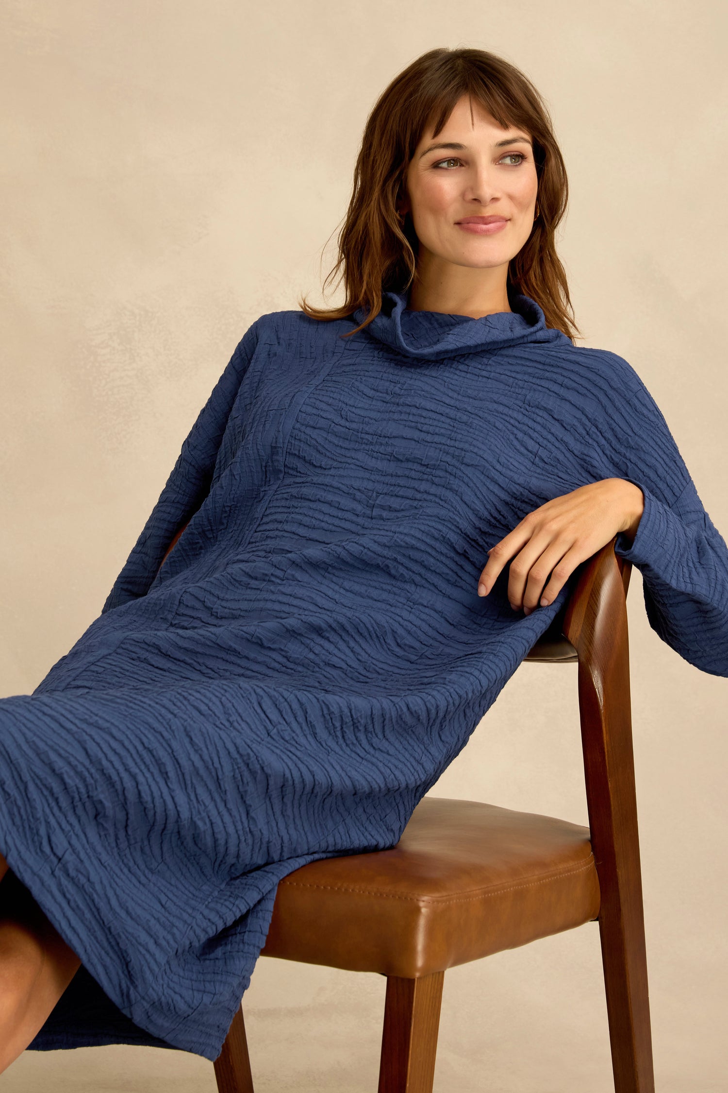 A woman wearing the Wave Textured Crinkle Dress, featuring a subtle bubble silhouette in vibrant cobalt, sits sideways on a brown chair, smiling as she gazes off to the side against a neutral background.