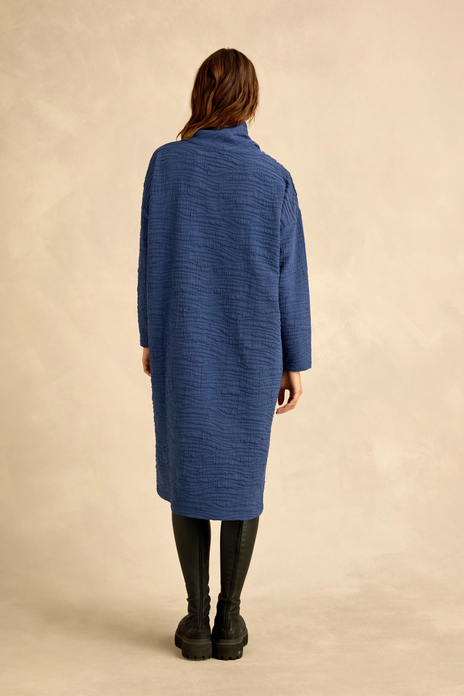 A person stands facing away, wearing the Wave Textured Crinkle Dress in blue—a knee-length piece with a subtle bubble silhouette—paired with black leggings and black boots, against a neutral beige background.