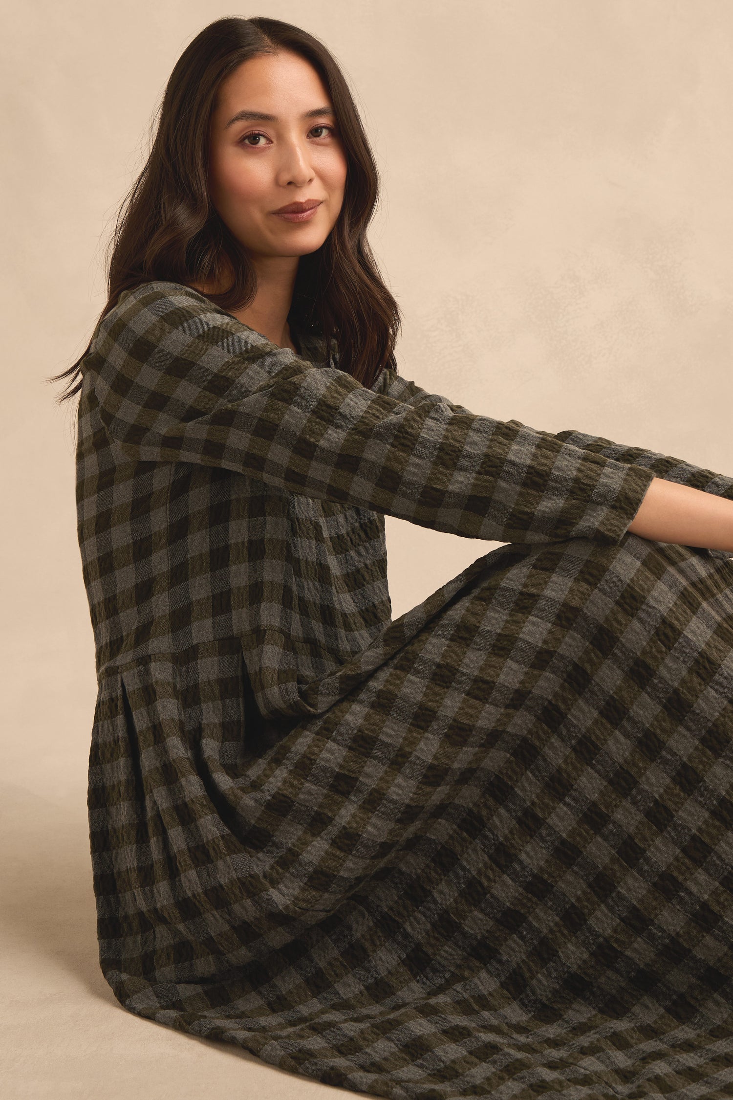A woman with long dark hair sits on the floor wearing the Crinkle Check Flared Dress, featuring a black and gray checkered pattern, looking at the camera with a slight smile against a neutral background.