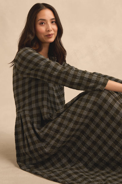 A woman with long dark hair sits on the floor wearing the Crinkle Check Flared Dress, featuring a black and gray checkered pattern, looking at the camera with a slight smile against a neutral background.