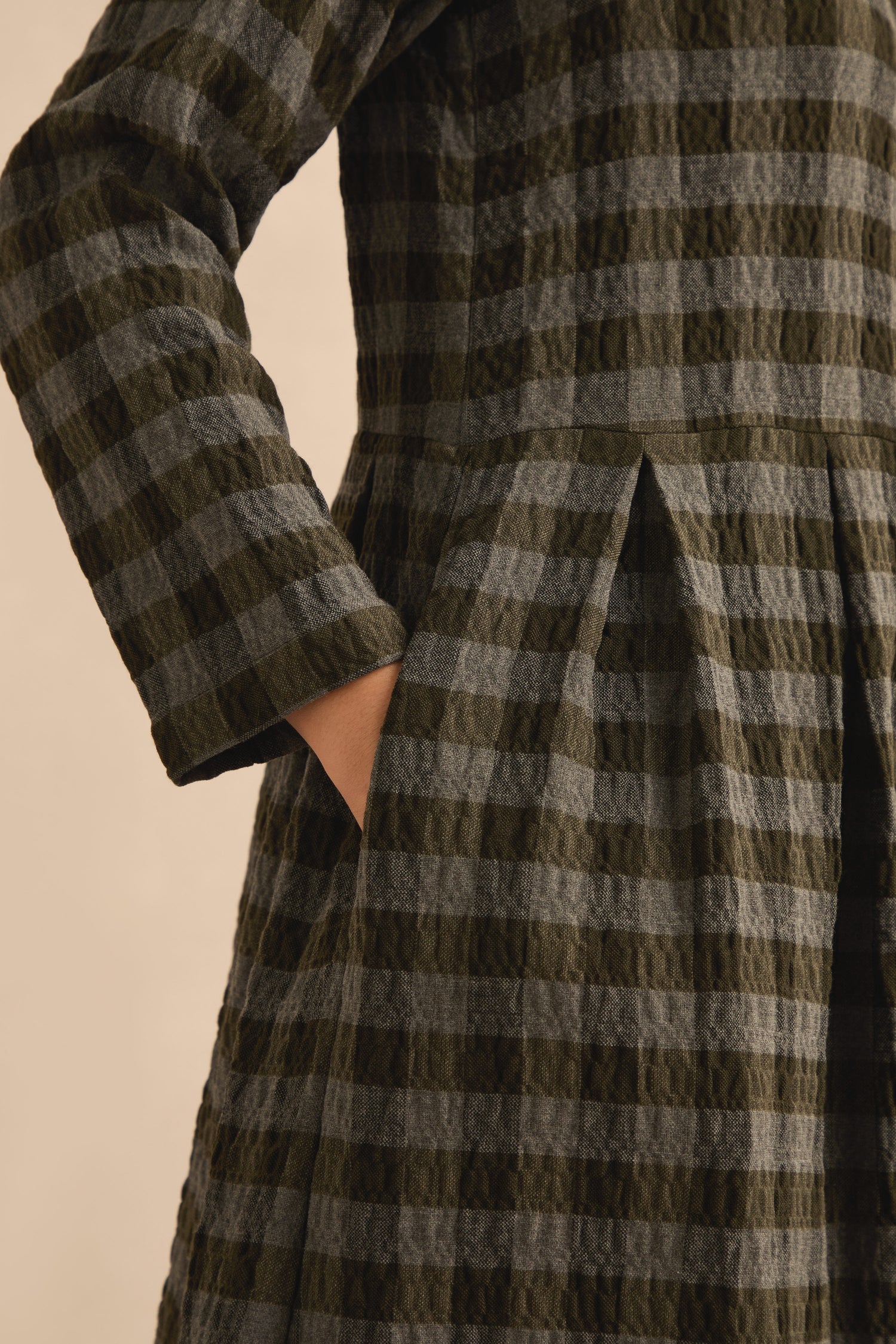 A person wearing the Crinkle Check Flared Dress, a long-sleeved, khaki-grey design with an A-line midi silhouette and one hand in the pocket.