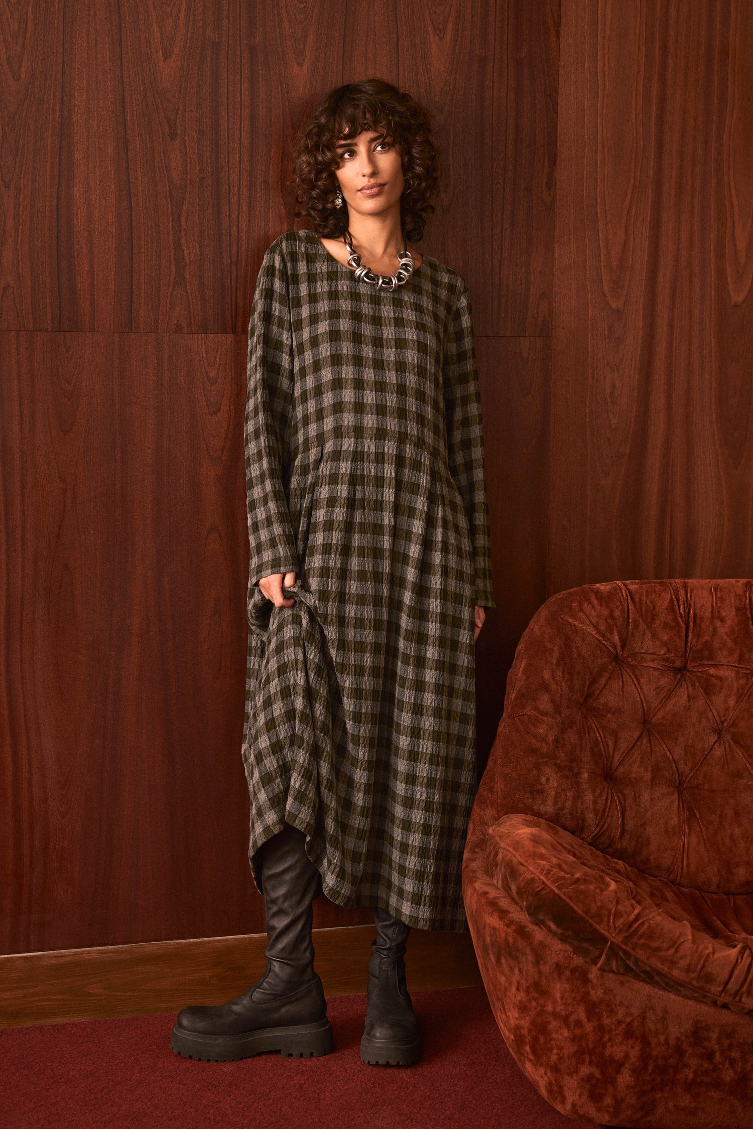 A person with curly hair stands by a wood-paneled wall wearing the Crinkle Check Flared Dress, chunky black boots, and a necklace, next to a brown upholstered chair on a red carpet.