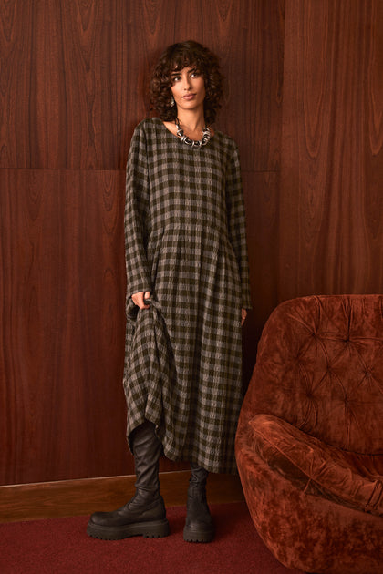 A person with curly hair stands by a wood-paneled wall wearing the Crinkle Check Flared Dress, chunky black boots, and a necklace, next to a brown upholstered chair on a red carpet.