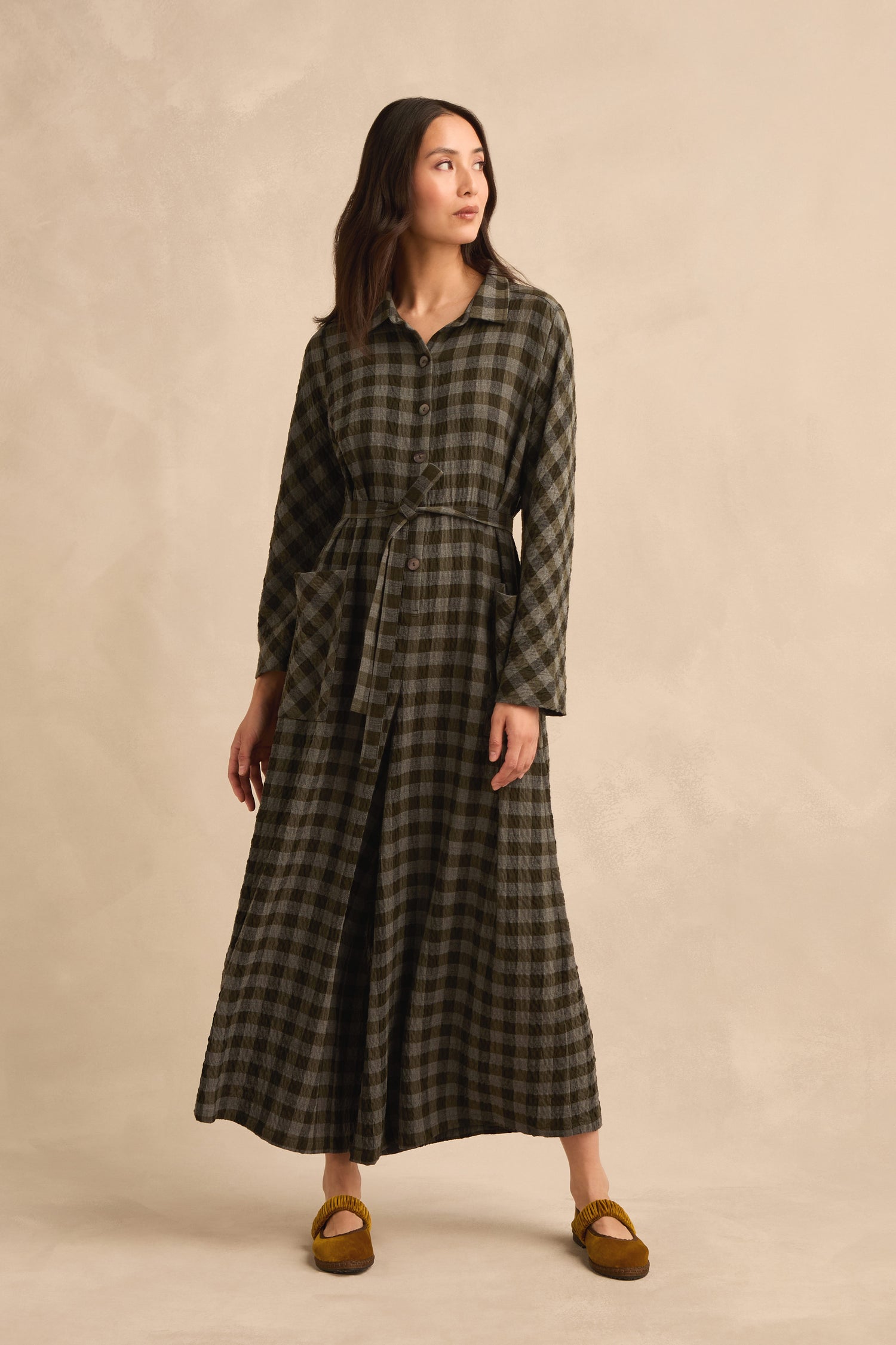 Woman wearing the Crinkle Check Flared Jumpsuit with a waist tie and brown slippers, standing against a beige background—her look echoing the effortless charm of relaxed, flared silhouettes.