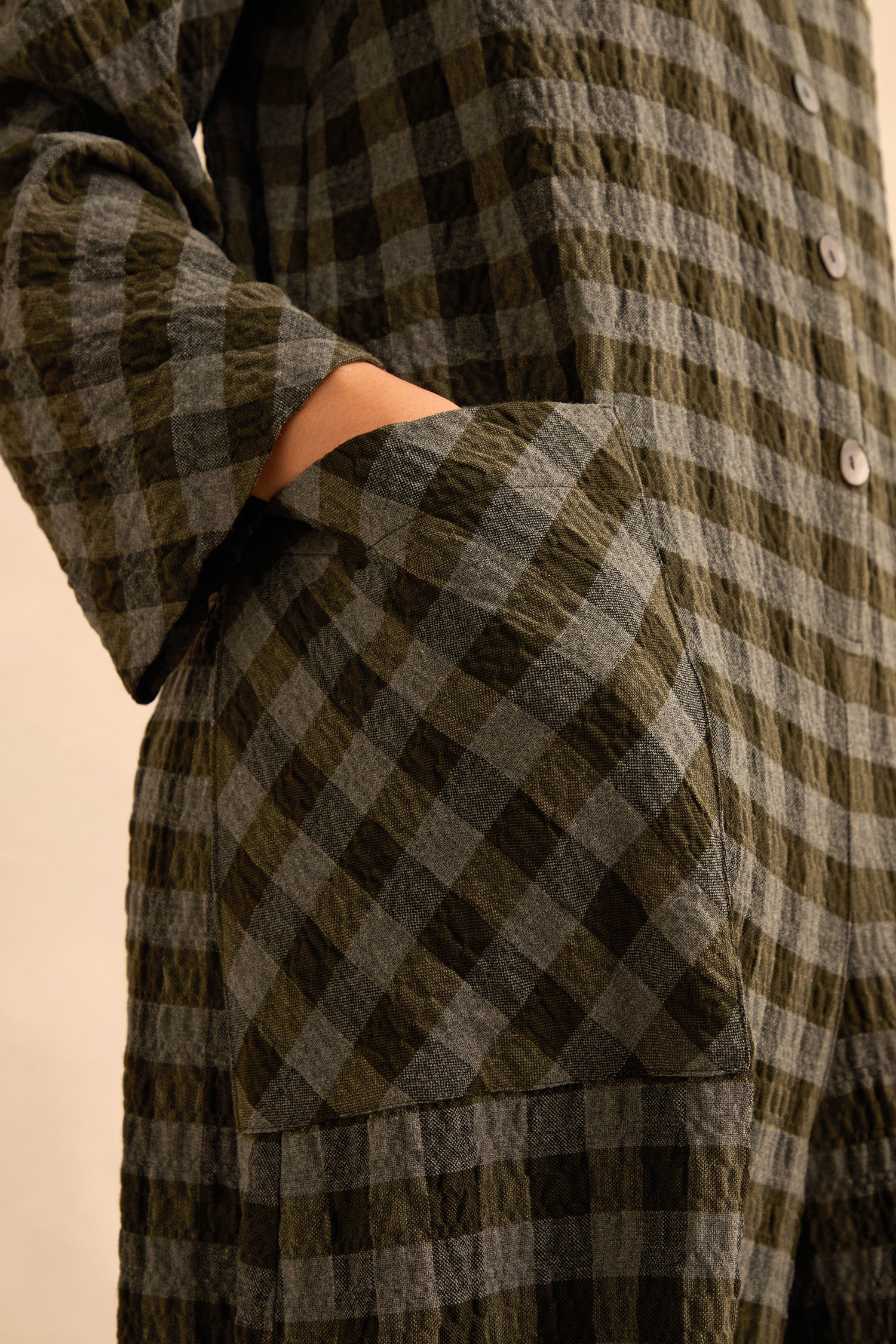 Close-up of a person wearing the Crinkle Check Flared Jumpsuit in green and gray plaid, featuring a large front pocket with a hand inside and visible buttons on the upper section.