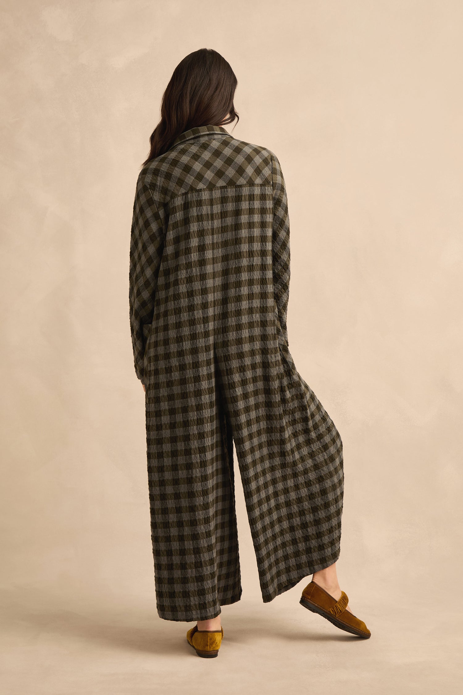 A person stands facing away, wearing the Crinkle Check Flared Jumpsuit with a relaxed fit and long plaid design, paired with mustard yellow shoes against a neutral background.