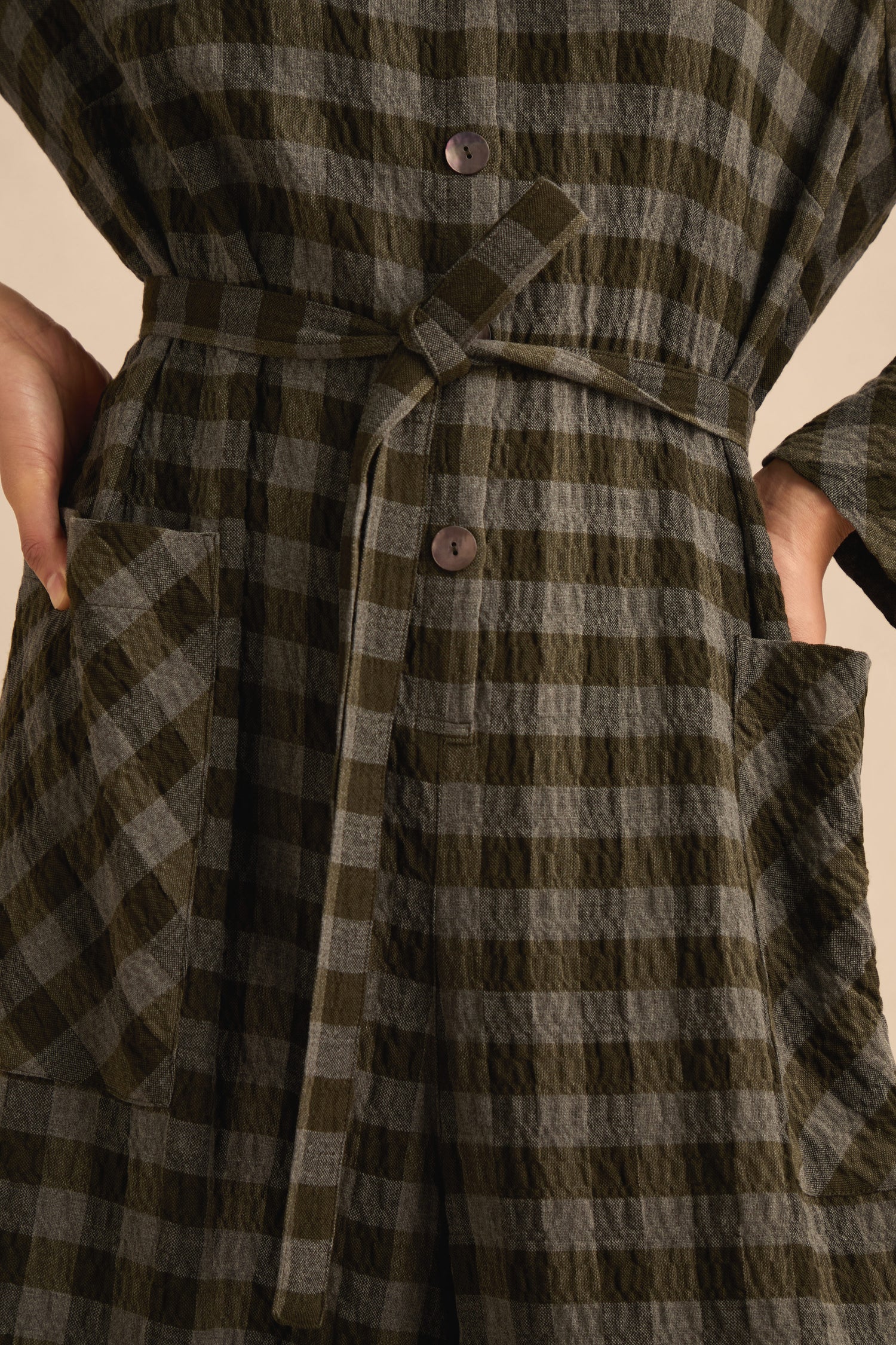 Close-up of a person wearing the Crinkle Check Flared Jumpsuit, which features a textured plaid button-down design, matching belt tied at the waist, and hands in front patch pockets.