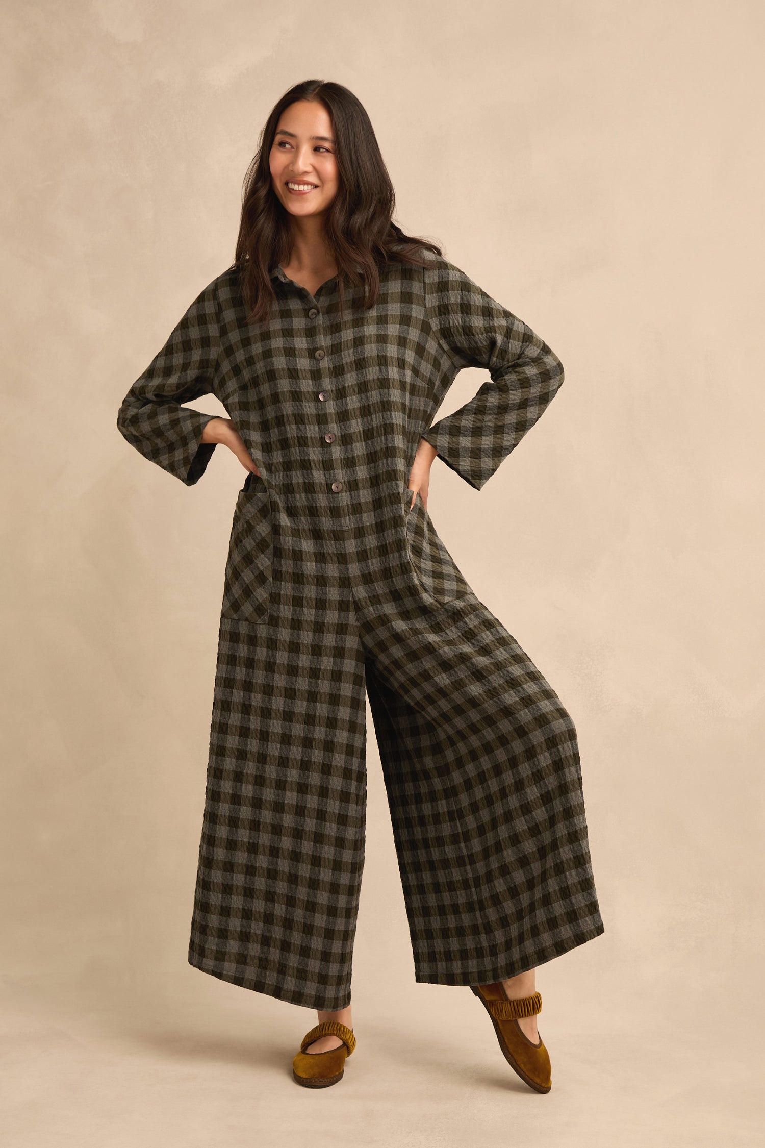A woman stands against a beige background with her hands in the pockets of a relaxed fit Crinkle Check Flared Jumpsuit, styled with wide legs and brown moccasins.