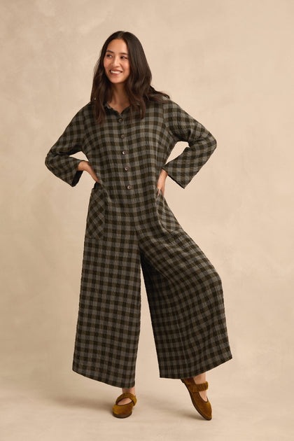 A woman stands against a beige background with her hands in the pockets of a relaxed fit Crinkle Check Flared Jumpsuit, styled with wide legs and brown moccasins.