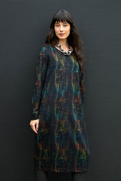 A woman wears a knee-length, long-sleeve Space Dye Jacquard Jersey Dress with a multicolored, textured vertical pattern on a black background, accessorized with a chunky, circular-link necklace.
