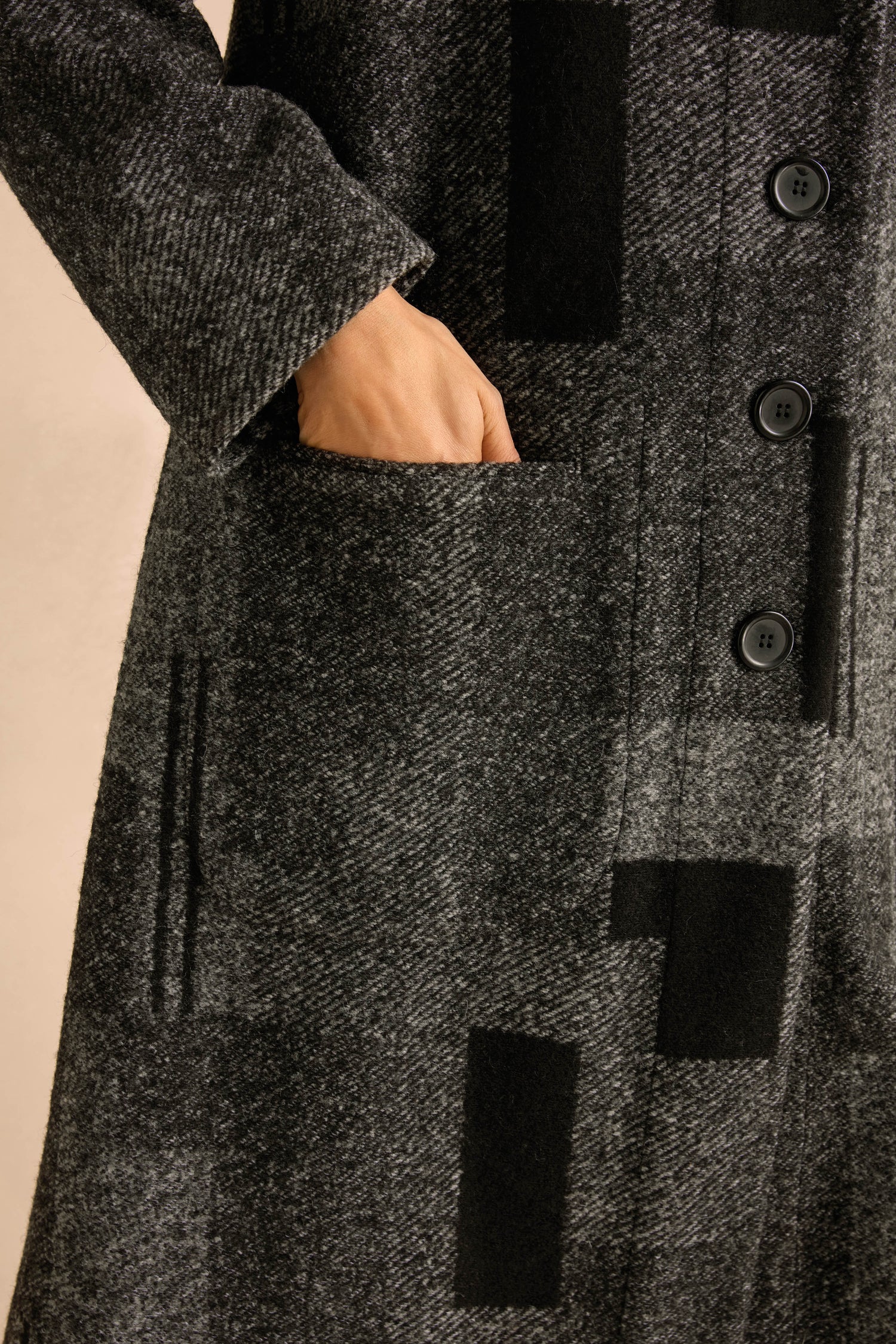 Close-up of a person’s hand in the pocket of the Melange Patchwork Wool Coat, featuring gray patterned wool with black buttons and geometric designs, highlighting its distinctive patchwork style.