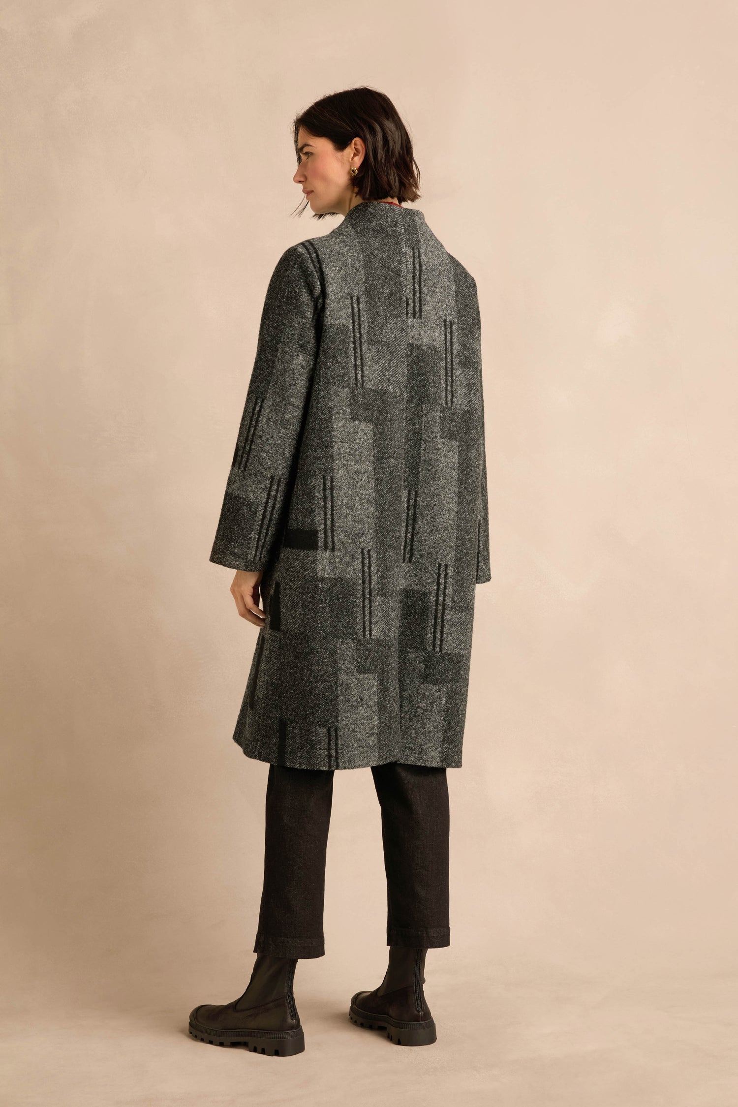 A person stands facing slightly away from the camera against a beige background, wearing the Melange Patchwork Wool Coat in gray, paired with black pants and black boots.
