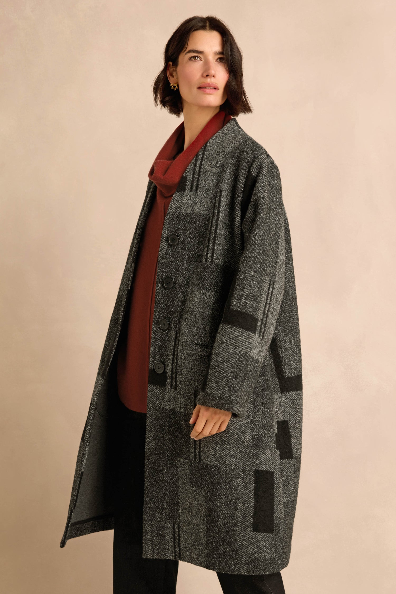 A woman wearing the Melange Patchwork Wool Coat—a long, gray, patterned wool coat—over a red top, stands against a beige background.