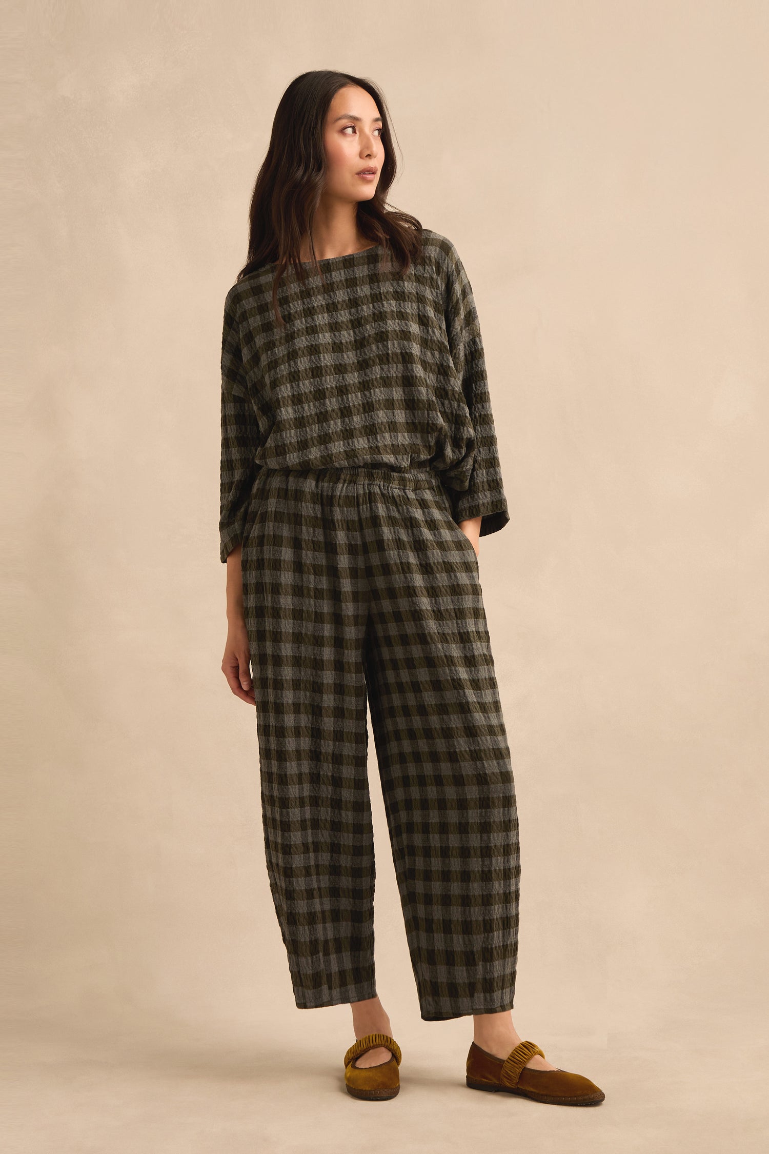 A woman stands against a beige background wearing a loose-fitting, checked long-sleeve top and the Crinkle Check Bubble Trouser in a cropped length, finished with mustard-colored loafers.