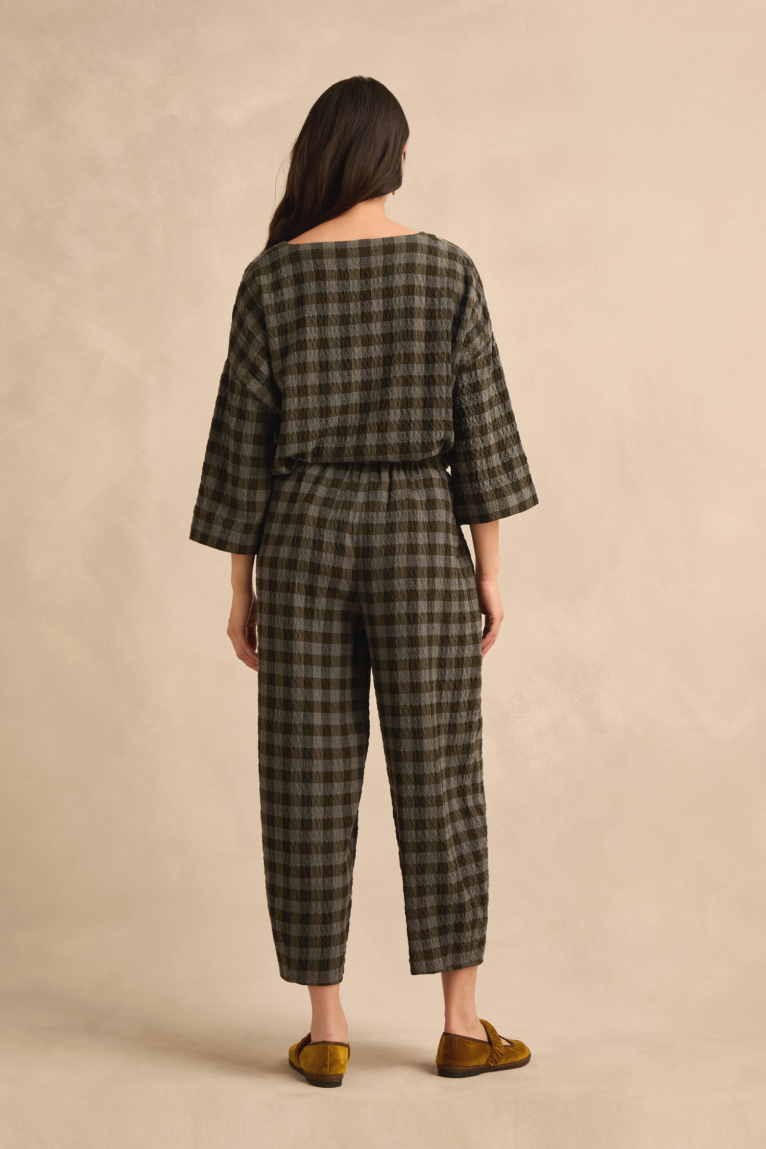 A woman stands facing away, wearing the Crinkle Check Bubble Trouser in a dark plaid pattern. The loose-fitting trousers feature a subtle bubble silhouette and a cropped, ankle-length cut. She pairs them with mustard yellow loafers against a beige background.