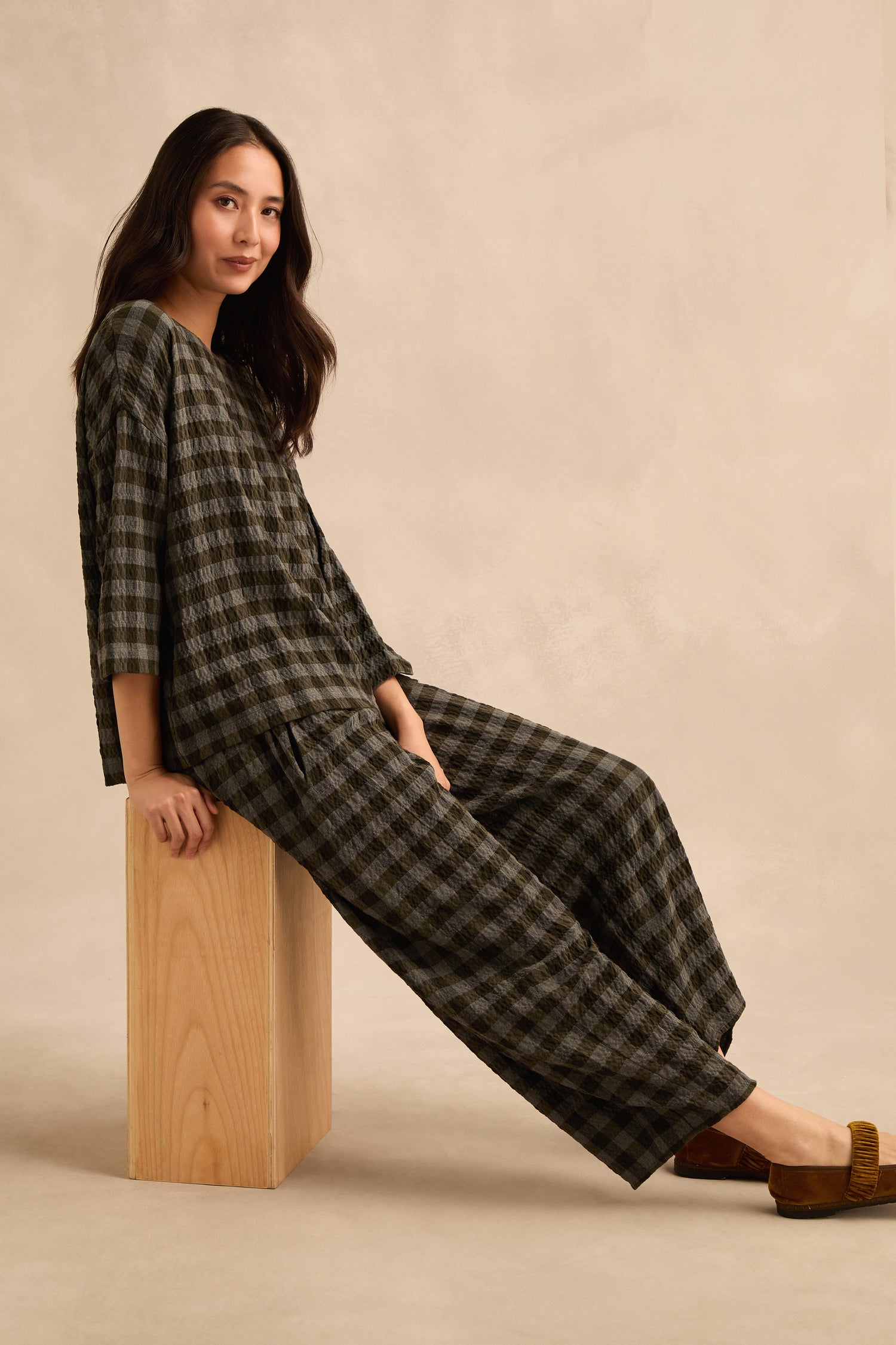 A woman seated on a wooden block, wearing a loose-fitting checked top and Crinkle Check Bubble Trouser with brown shoes, against a plain beige background.