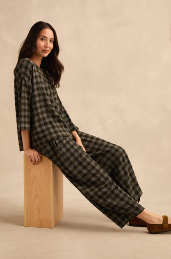 A woman seated on a wooden block, wearing a loose-fitting checked top and Crinkle Check Bubble Trouser with brown shoes, against a plain beige background.