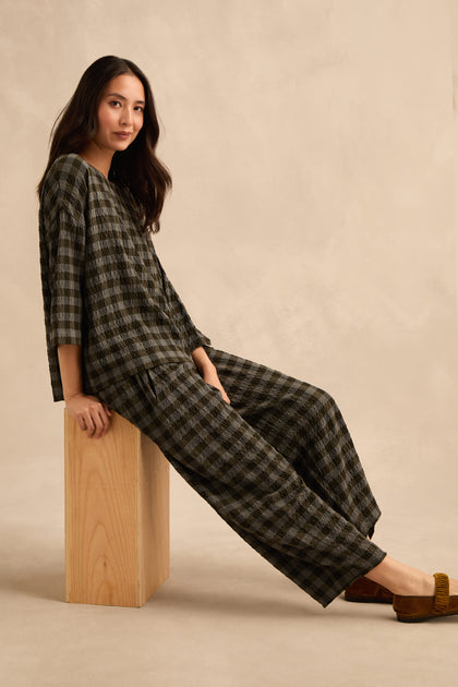 A woman seated on a wooden block, wearing a loose-fitting checked top and Crinkle Check Bubble Trouser with brown shoes, against a plain beige background.