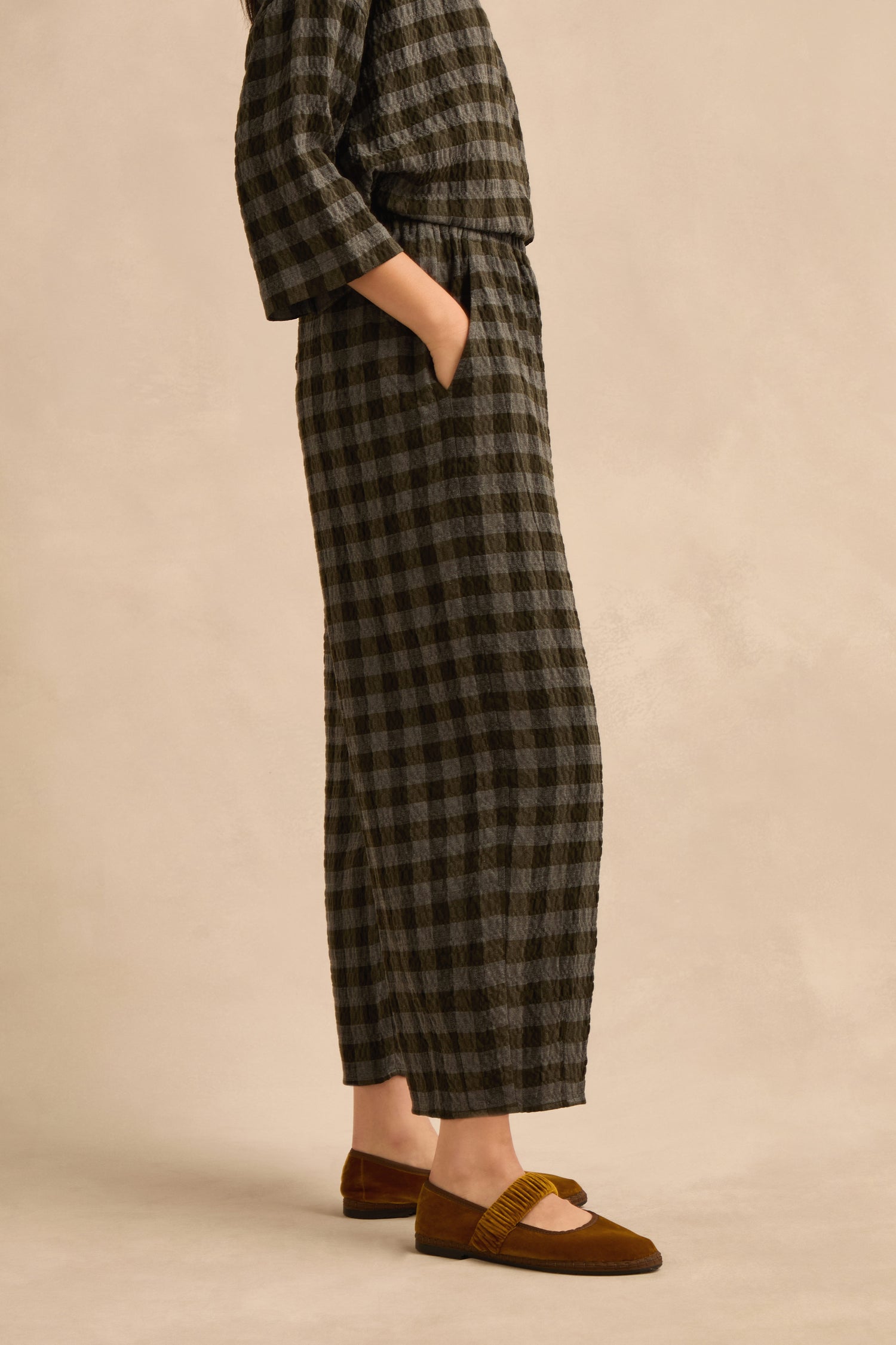 A person wearing a long-sleeved, ankle-length black and gray checkered dress with hands in pockets and brown flats stands against a neutral background—the subtly voluminous bubble silhouette evokes the modern style of the Crinkle Check Bubble Trouser.