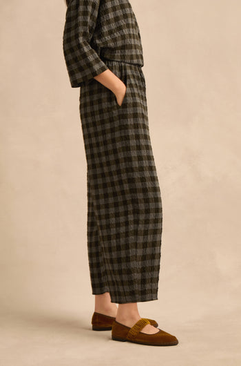 A person wearing a long-sleeved, ankle-length black and gray checkered dress with hands in pockets and brown flats stands against a neutral background—the subtly voluminous bubble silhouette evokes the modern style of the Crinkle Check Bubble Trouser.
