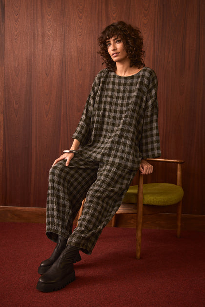A person with curly hair sits on a wooden chair in a room with wood-paneled walls and red carpet, wearing Crinkle Check Bubble Trousers with a cropped leg and black platform boots.