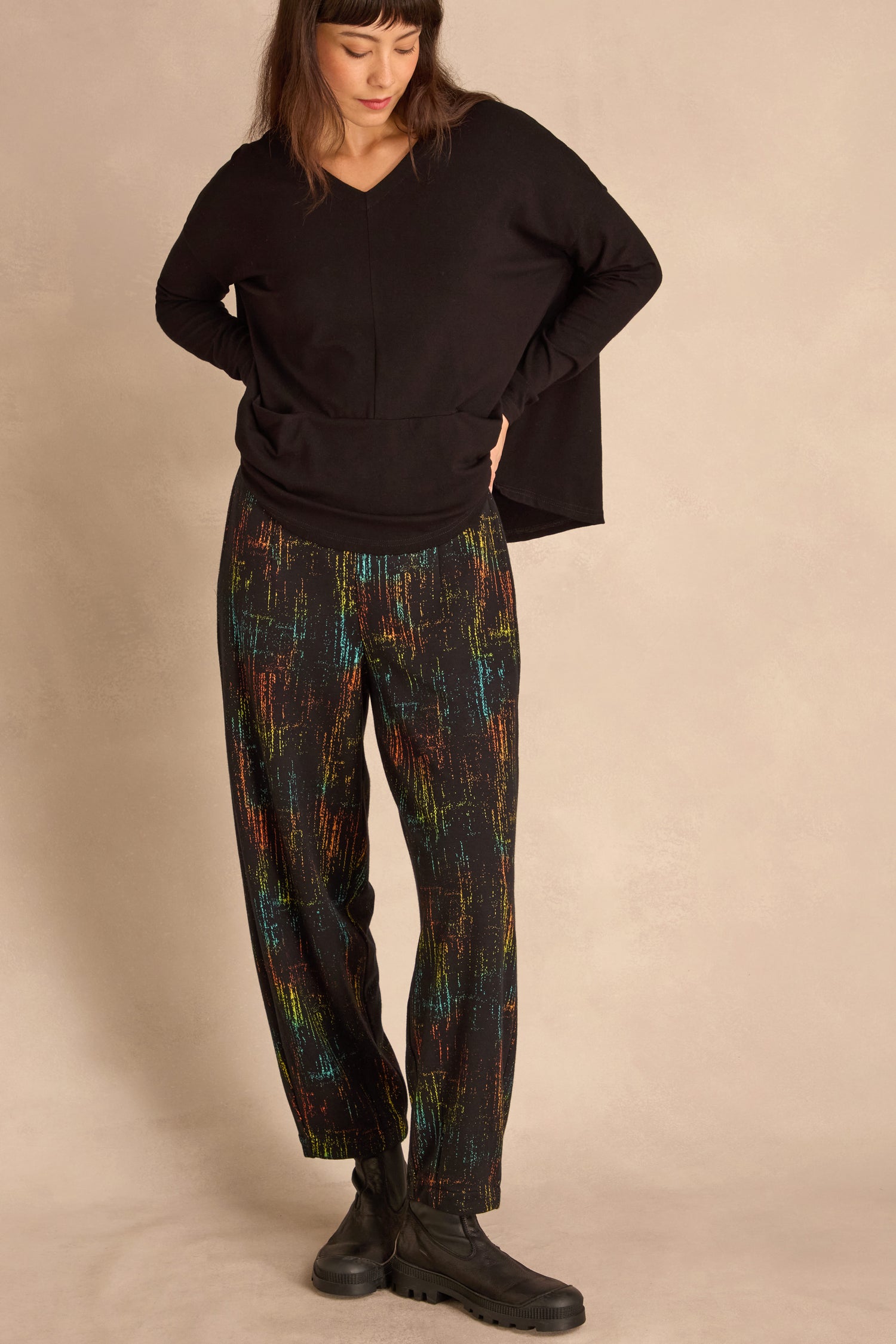 Woman wearing a loose black top, Space Dye Jacquard Bubble Trousers, and black boots stands against a neutral background, looking down with one hand on her hip.