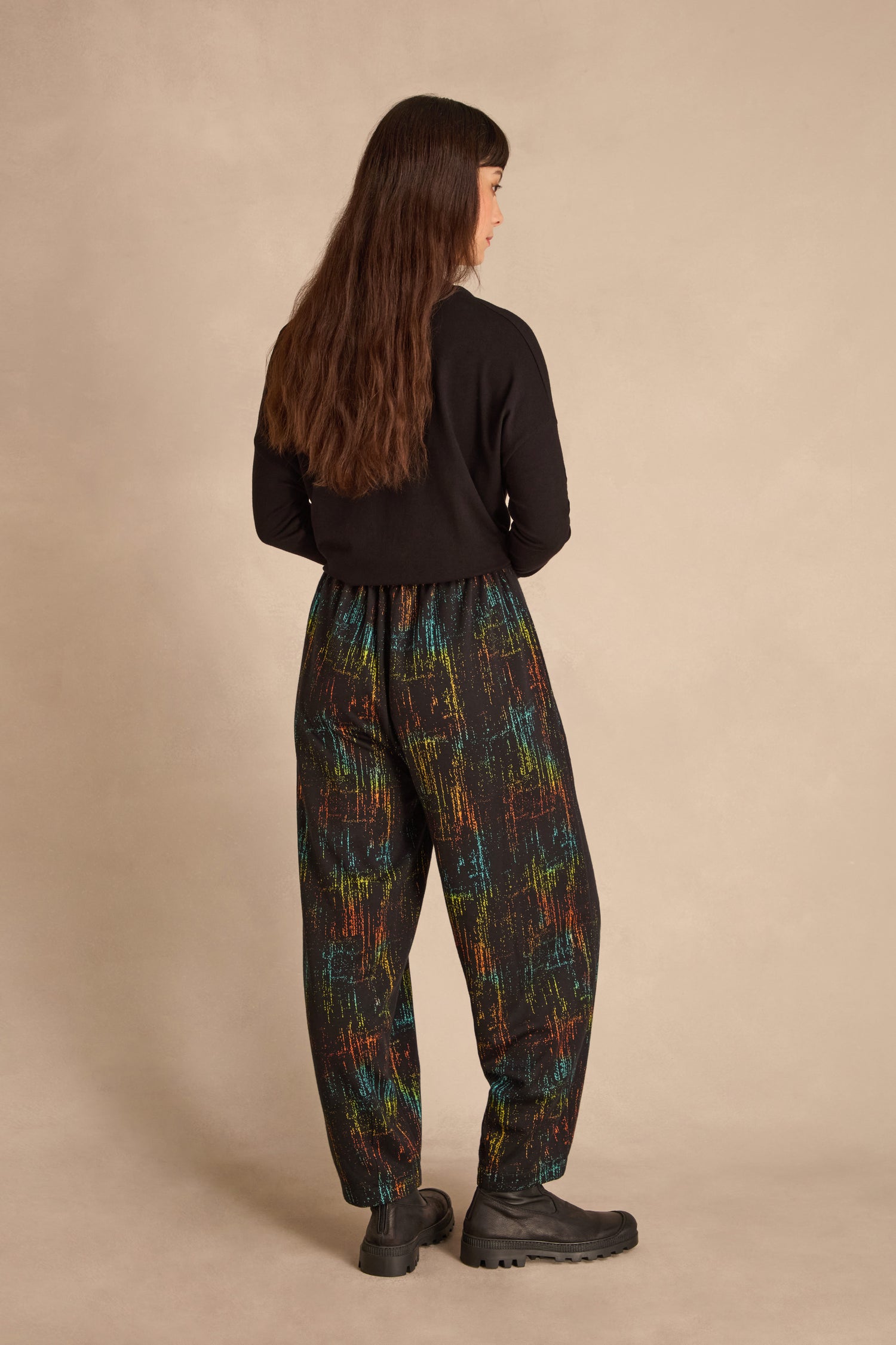 Person with long brown hair stands facing away, wearing black jacquard jersey top, Space Dye Jacquard Bubble Trousers, and black boots against a neutral background.