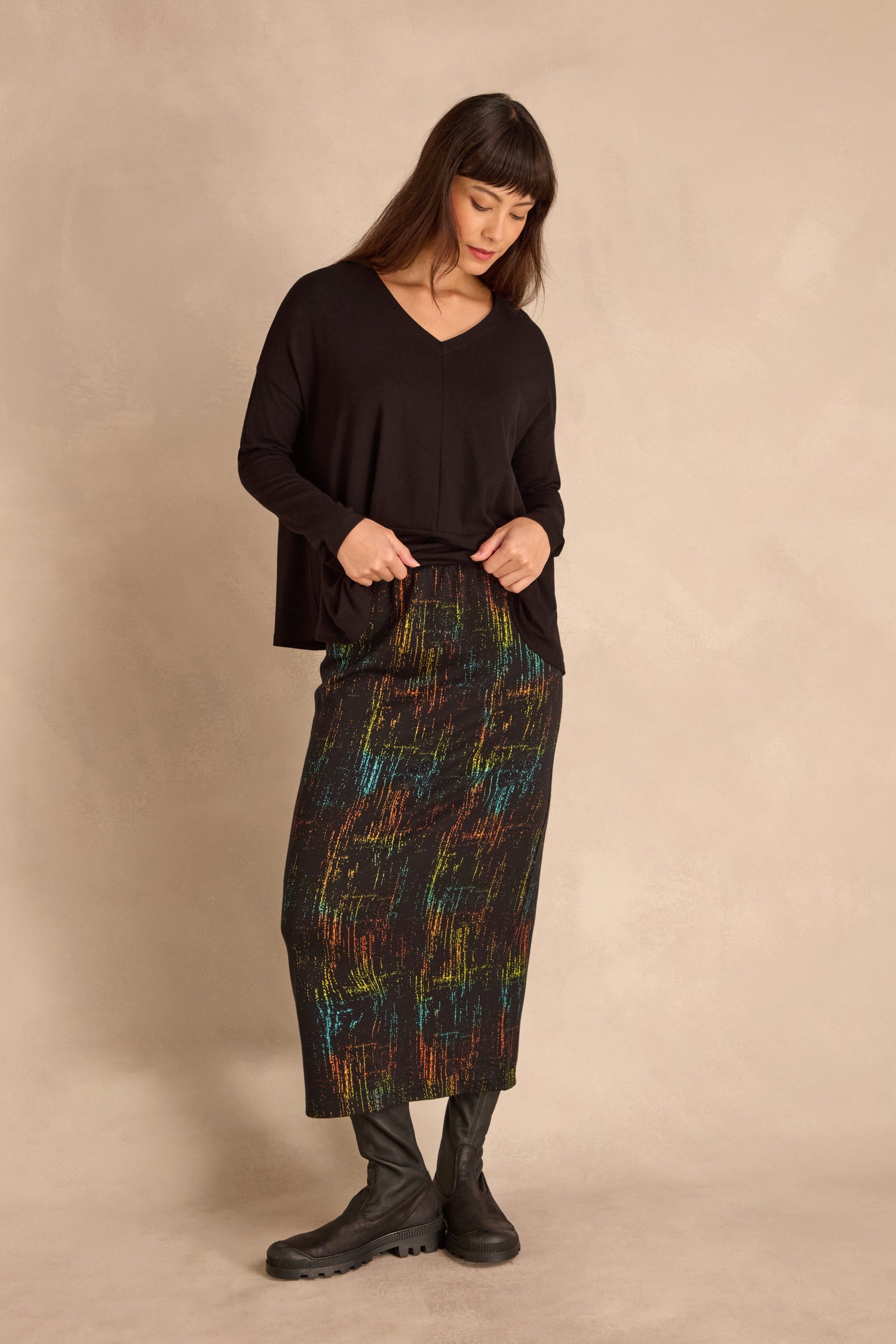 Woman wearing a loose black top, the Space Dye Jacquard Tube Skirt with colorful abstract patterns, and black boots, standing against a beige background.