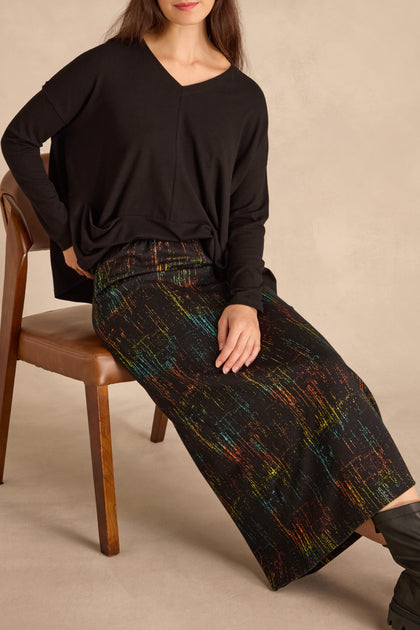 Woman seated on a wooden chair wearing a loose black long-sleeve top and the Space Dye Jacquard Tube Skirt with colorful abstract patterns.
