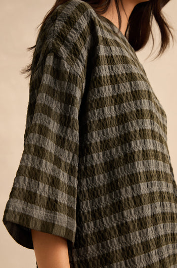 A person wearing the Crinkle Check Boxy Top—a textured, short-sleeved shirt in green and gray stripes with a boxy silhouette—shown from the shoulder down against a neutral background.