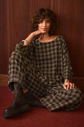A person with curly hair sits on a red carpet, styled in the Crinkle Check Boxy Top and black boots, posed against a wooden wall.