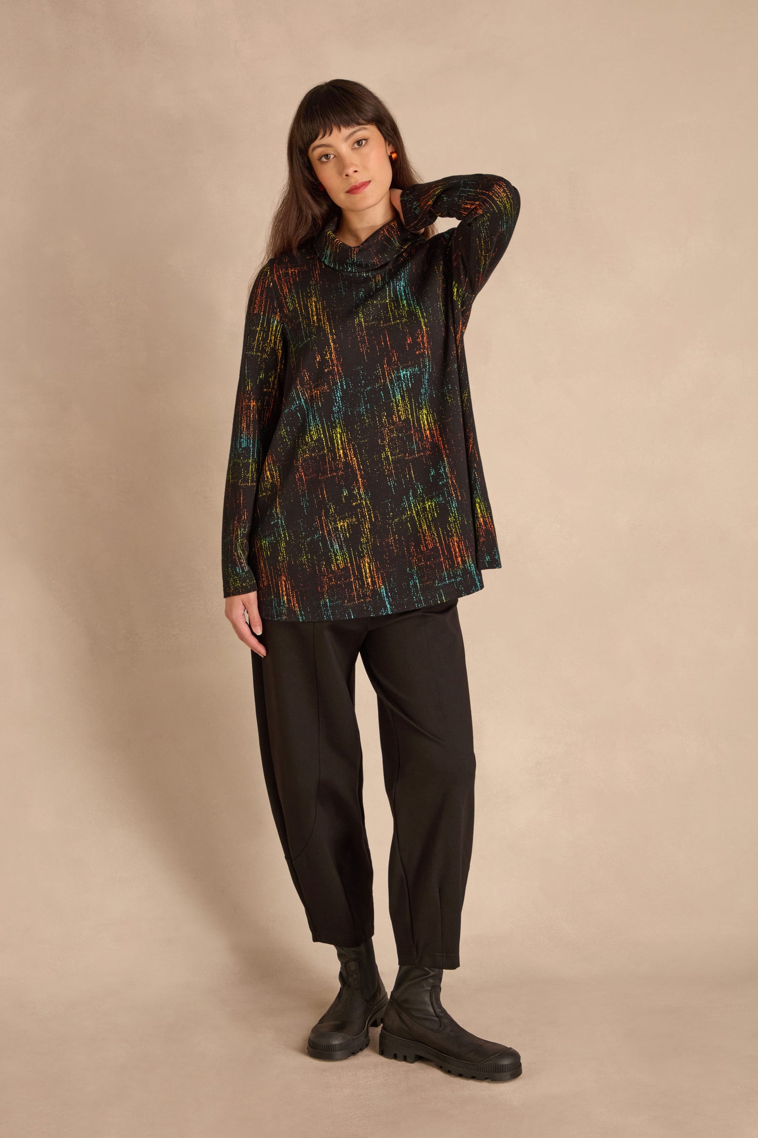 A woman stands against a plain background in the Space Dye Jacquard Jersey Tunic, paired with loose black pants and boots, her left hand touching her head.