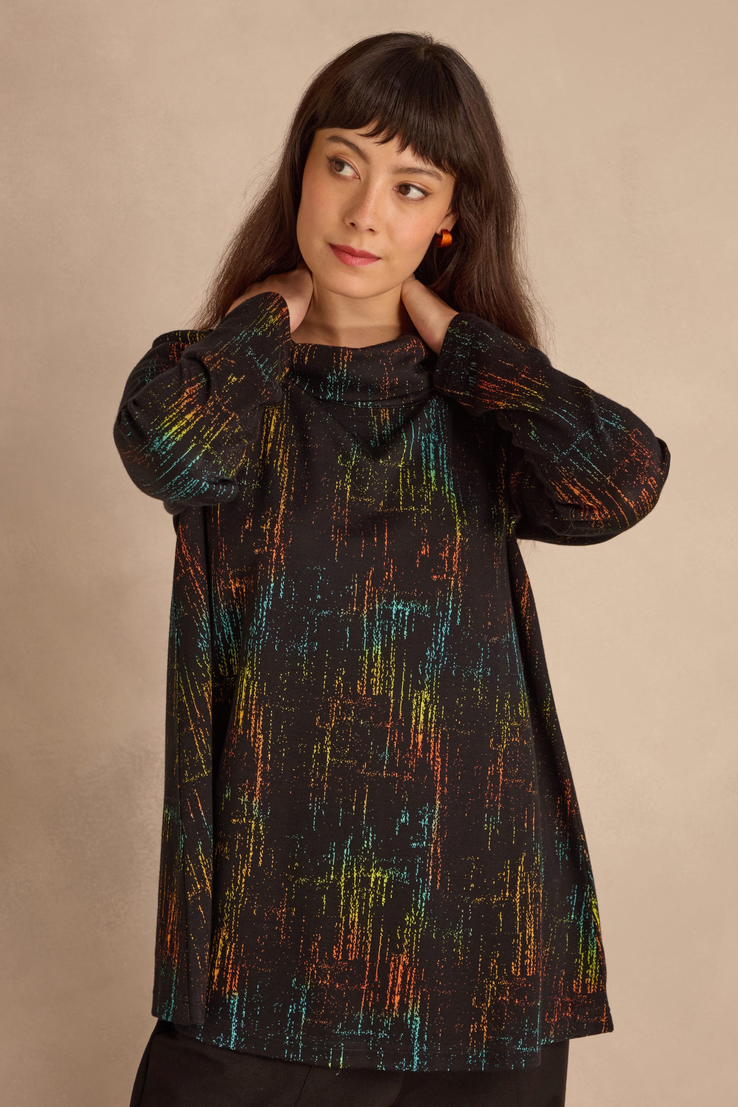 A woman with long brown hair models the Space Dye Jacquard Jersey Tunic, featuring colorful streaks, standing against a neutral background with hands near her neck.