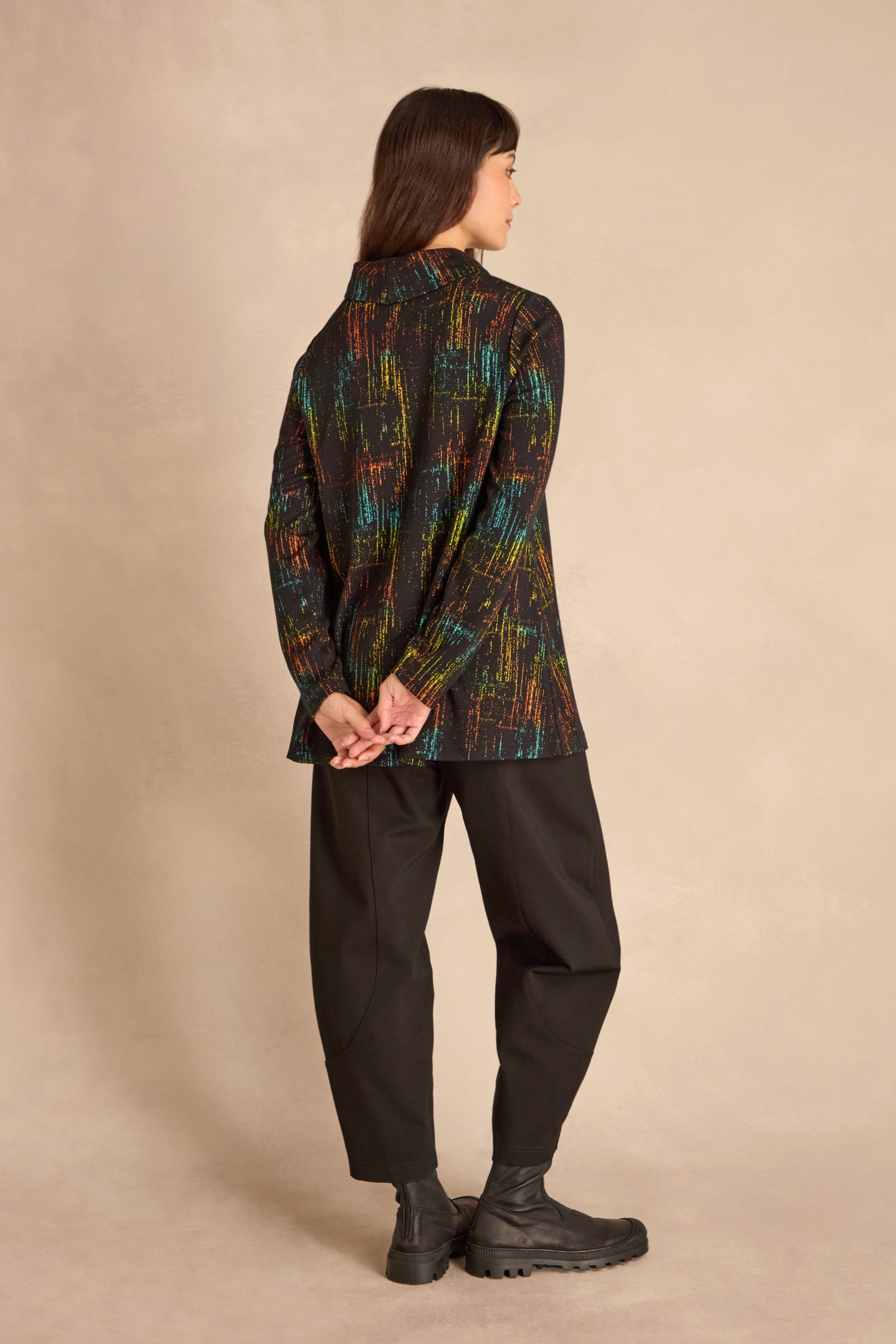 A woman stands facing away, hands clasped behind her back, wearing the Space Dye Jacquard Jersey Tunic with black pants and boots against a beige background.