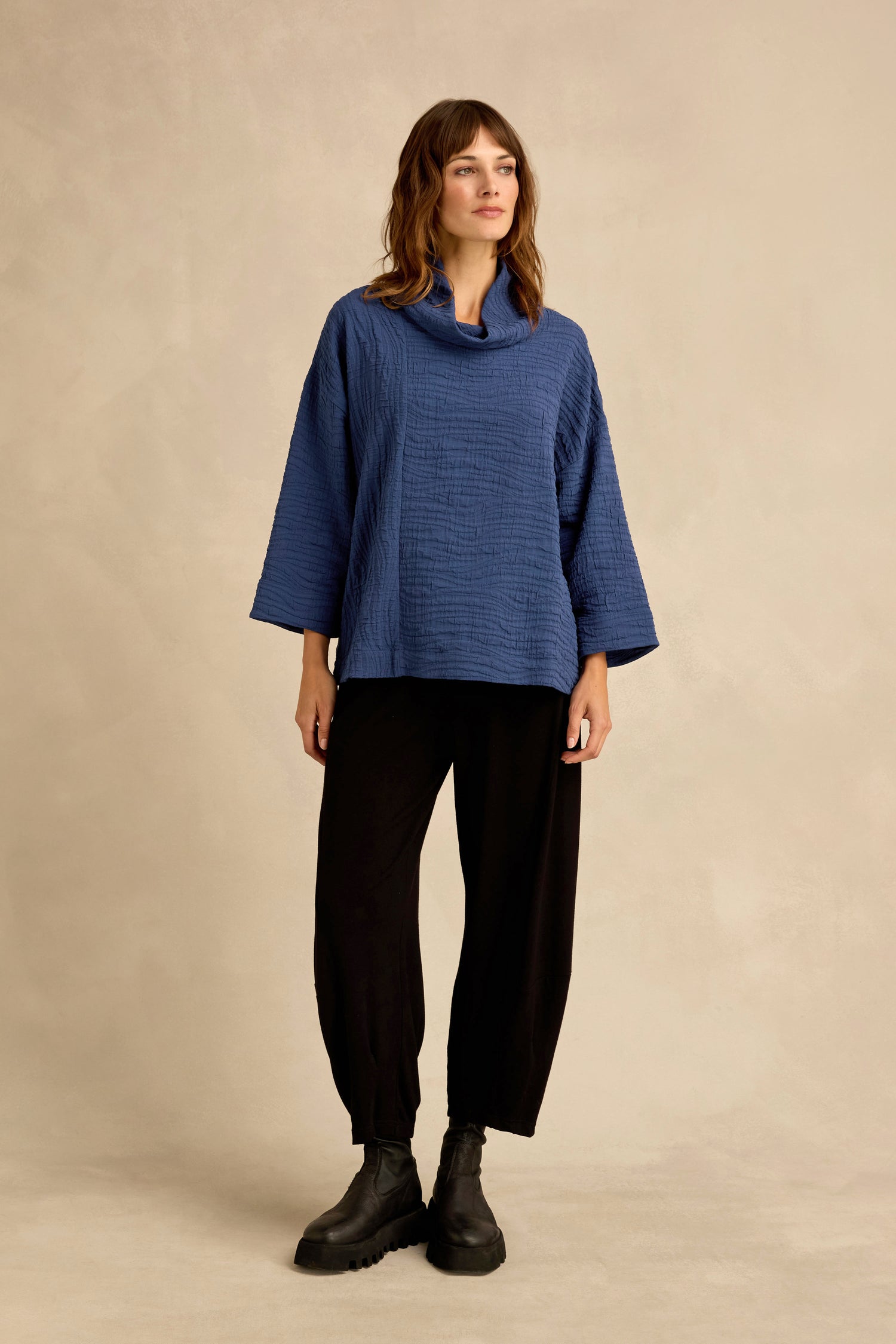A woman stands against a beige background, wearing the Cobalt Wave Textured Crinkle Cowl Neck Top in a relaxed boxy silhouette, paired with black pants and black boots.
