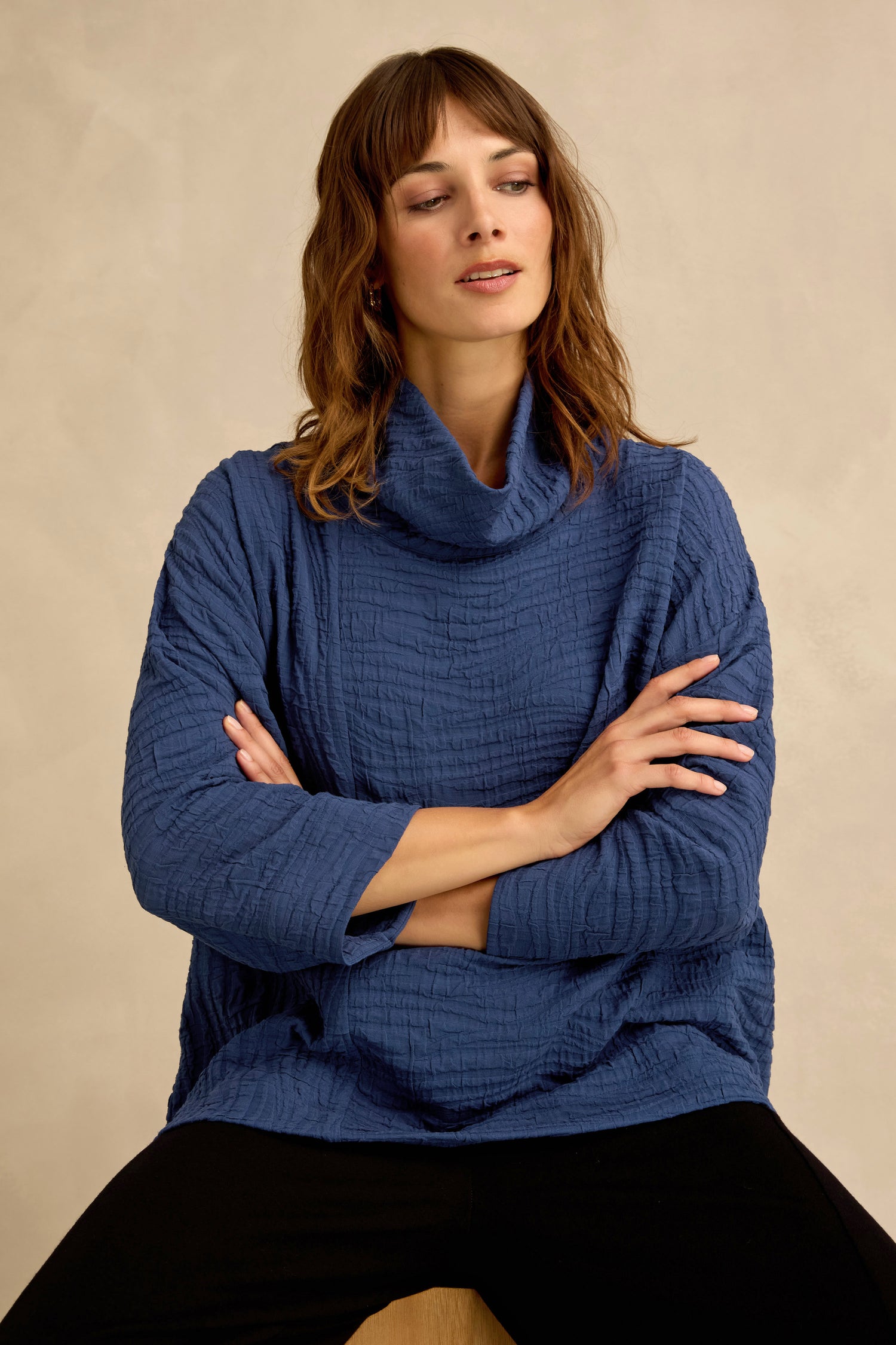 A woman sits with her arms crossed, wearing the Cobalt Wave Textured Crinkle Cowl Neck Top paired with black pants, set against a plain beige background.