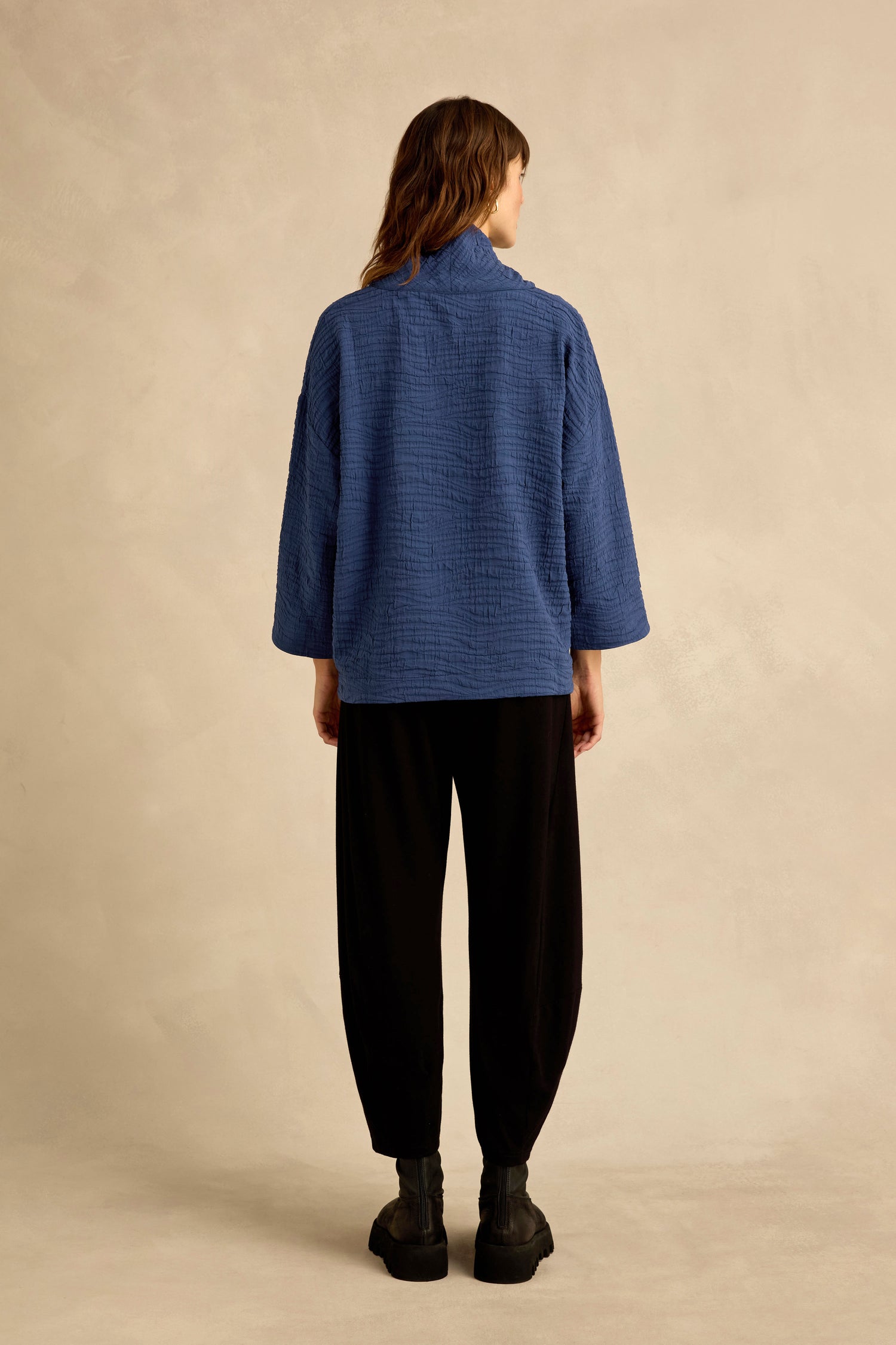 Person standing against a beige background, facing away, wearing the Cobalt Wave Textured Crinkle Cowl Neck Top in a relaxed boxy shape, paired with black pants and black boots.