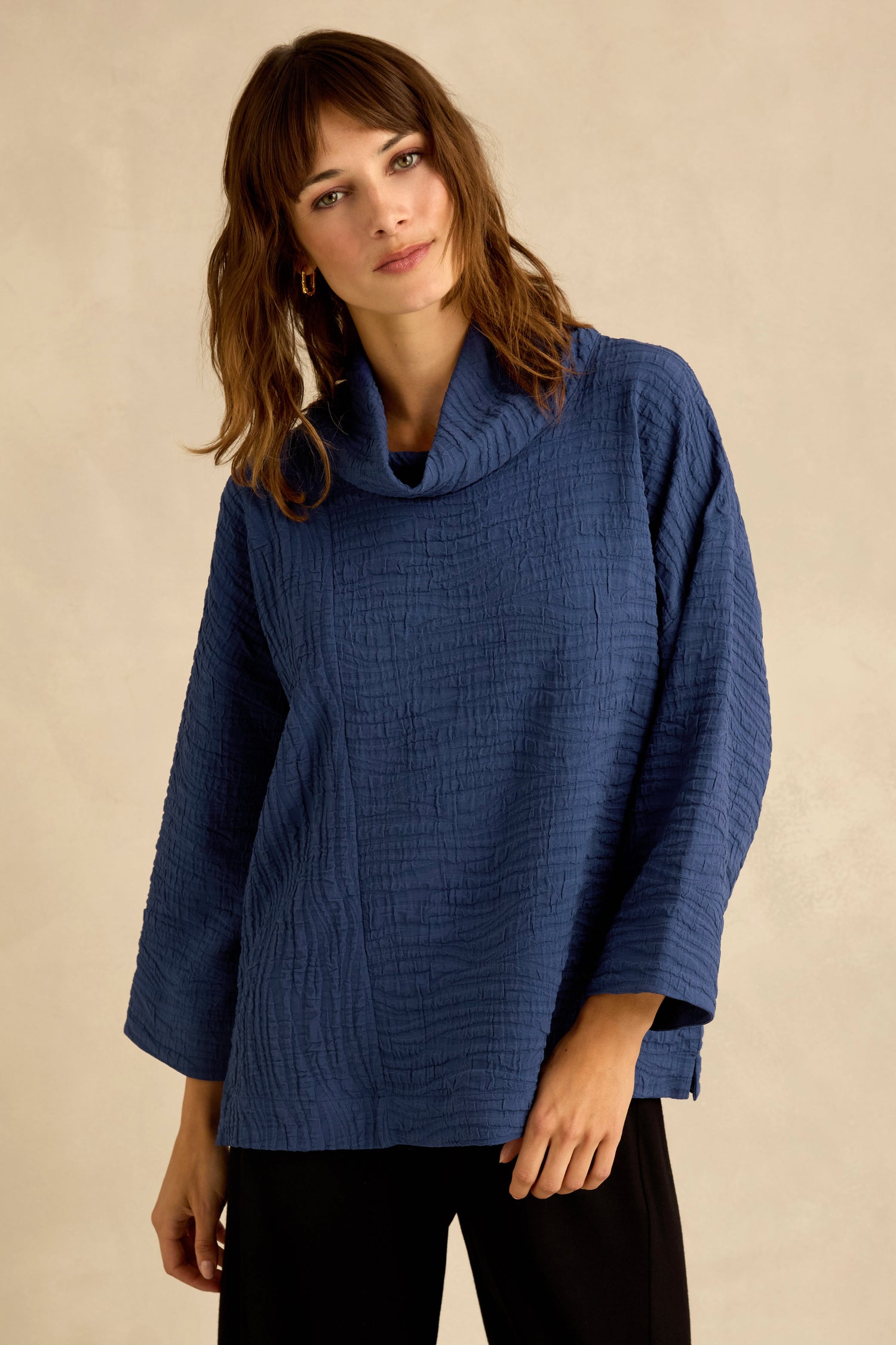 A woman is wearing the Cobalt Wave Textured Crinkle Cowl Neck Top with long sleeves and black pants, standing against a plain beige background. The top’s relaxed boxy shape adds effortless style to her look.