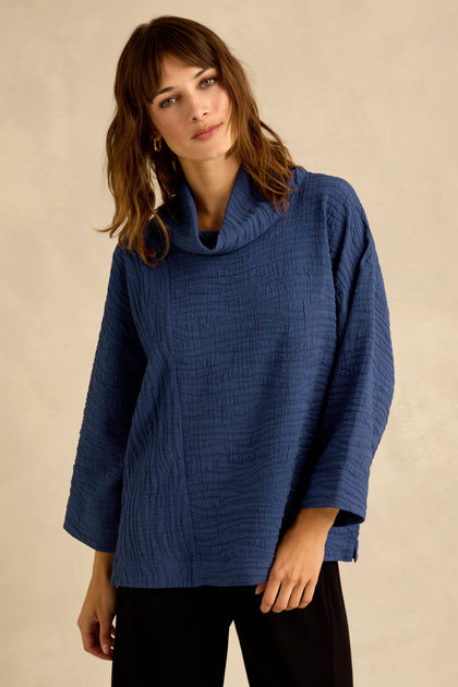 A woman is wearing the Cobalt Wave Textured Crinkle Cowl Neck Top with long sleeves and black pants, standing against a plain beige background. The top’s relaxed boxy shape adds effortless style to her look.