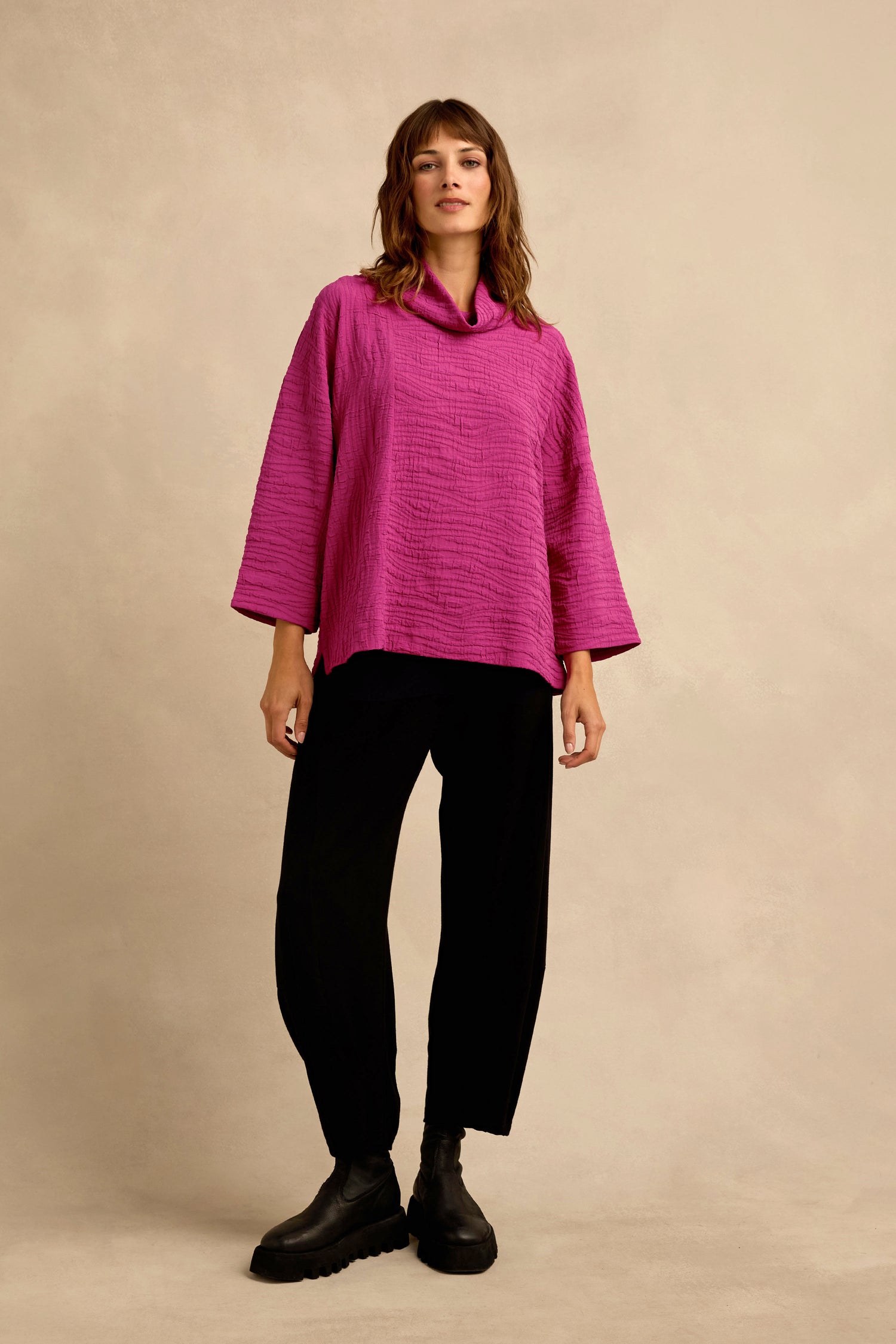 A woman stands against a beige background wearing the Magenta Wave Textured Crinkle Cowl Neck Top with wide sleeves, paired with black pants and black boots.