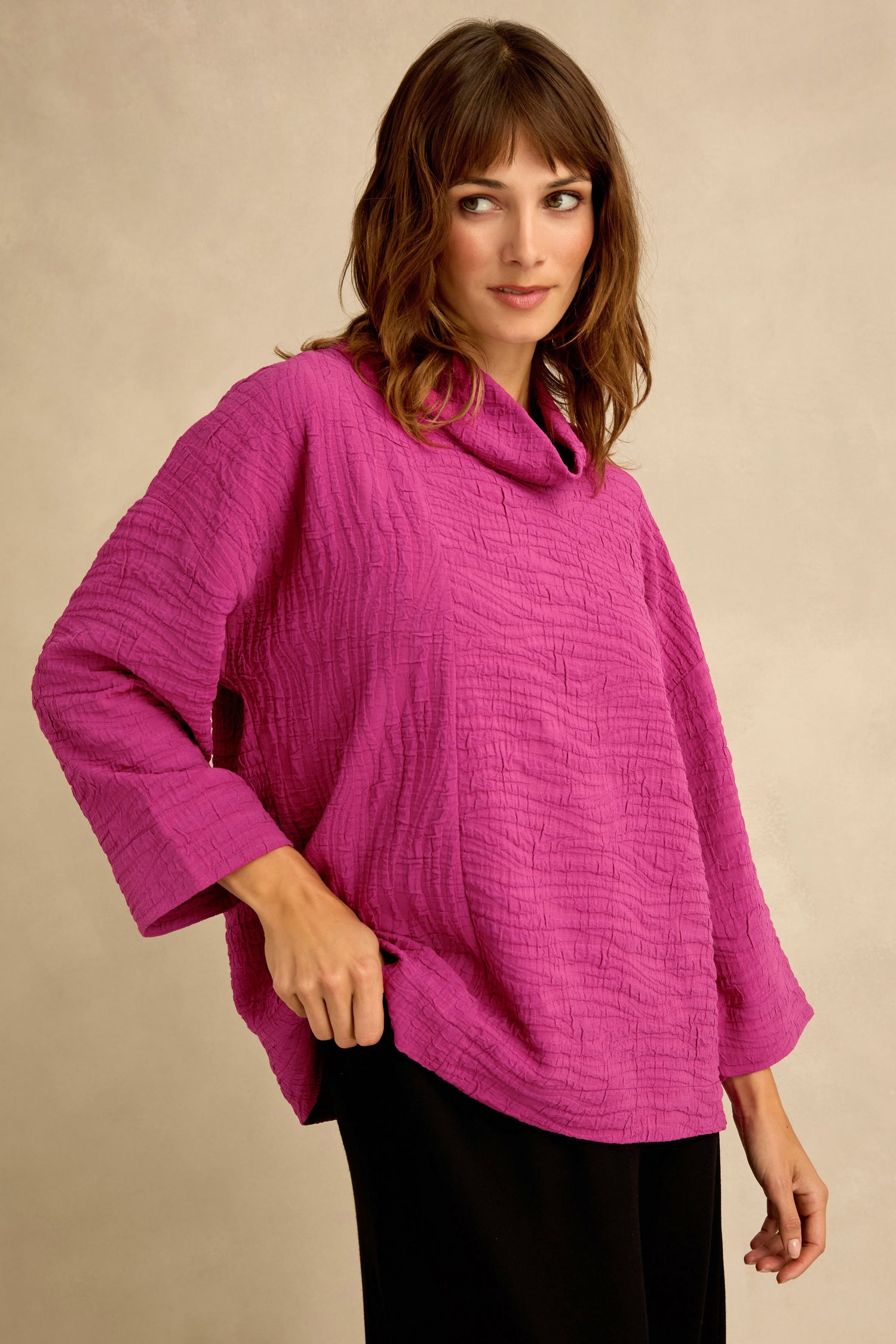 A woman stands against a neutral background wearing a Magenta Wave Textured Crinkle Cowl Neck Top with three-quarter sleeves and black pants.