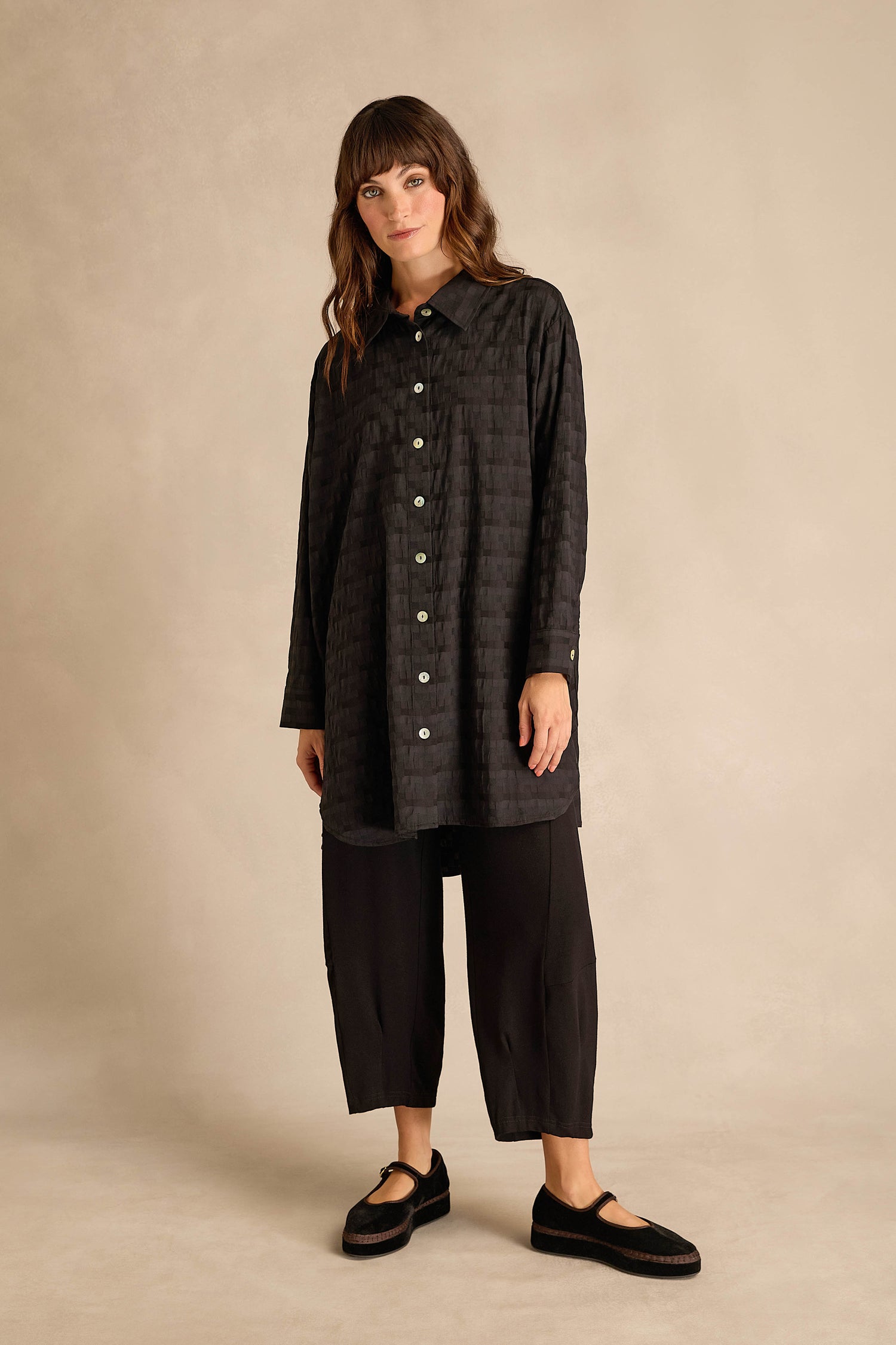 Woman stands against a beige background wearing the Jacquard Squares Long Line Shirt styled as a button-up, paired with loose black pants and black shoes. She has long brown hair and a neutral expression.