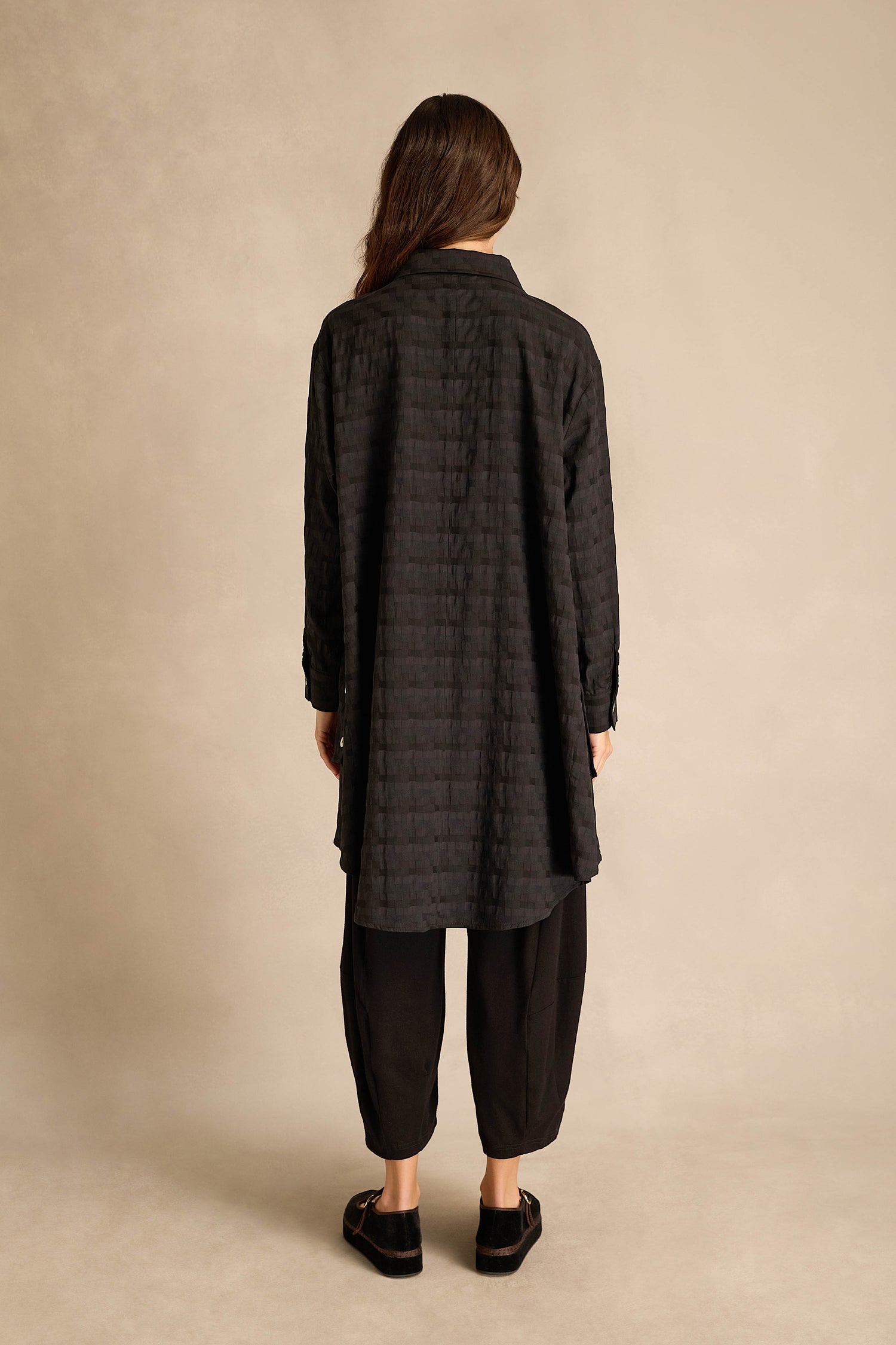A person stands with their back to the camera, wearing the Jacquard Squares Long Line Shirt—crafted from a cotton-viscose blend with a woven grid texture—paired with loose black pants and black shoes against a neutral background.