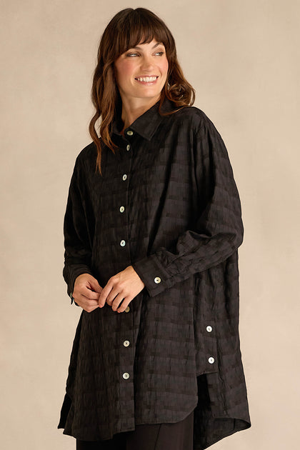 A woman smiles and looks sideways, wearing the Jacquard Squares Long Line Shirt—a textured black shirt featuring a subtle checkered pattern and white buttons—standing against a plain beige background.