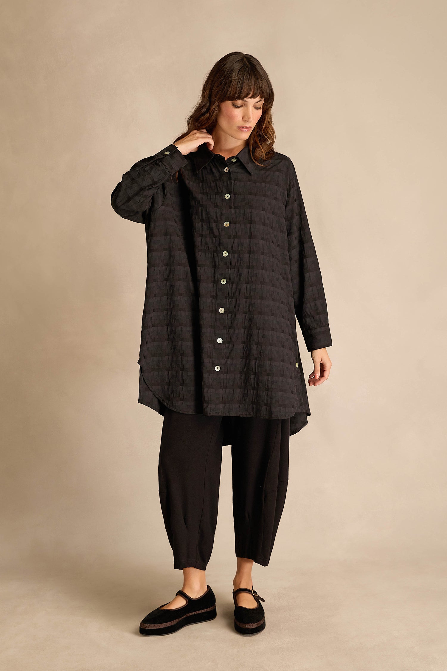 A woman wearing the Jacquard Squares Long Line Shirt in black, paired with black cropped pants, stands against a beige background, looking down and touching her hair with one hand.